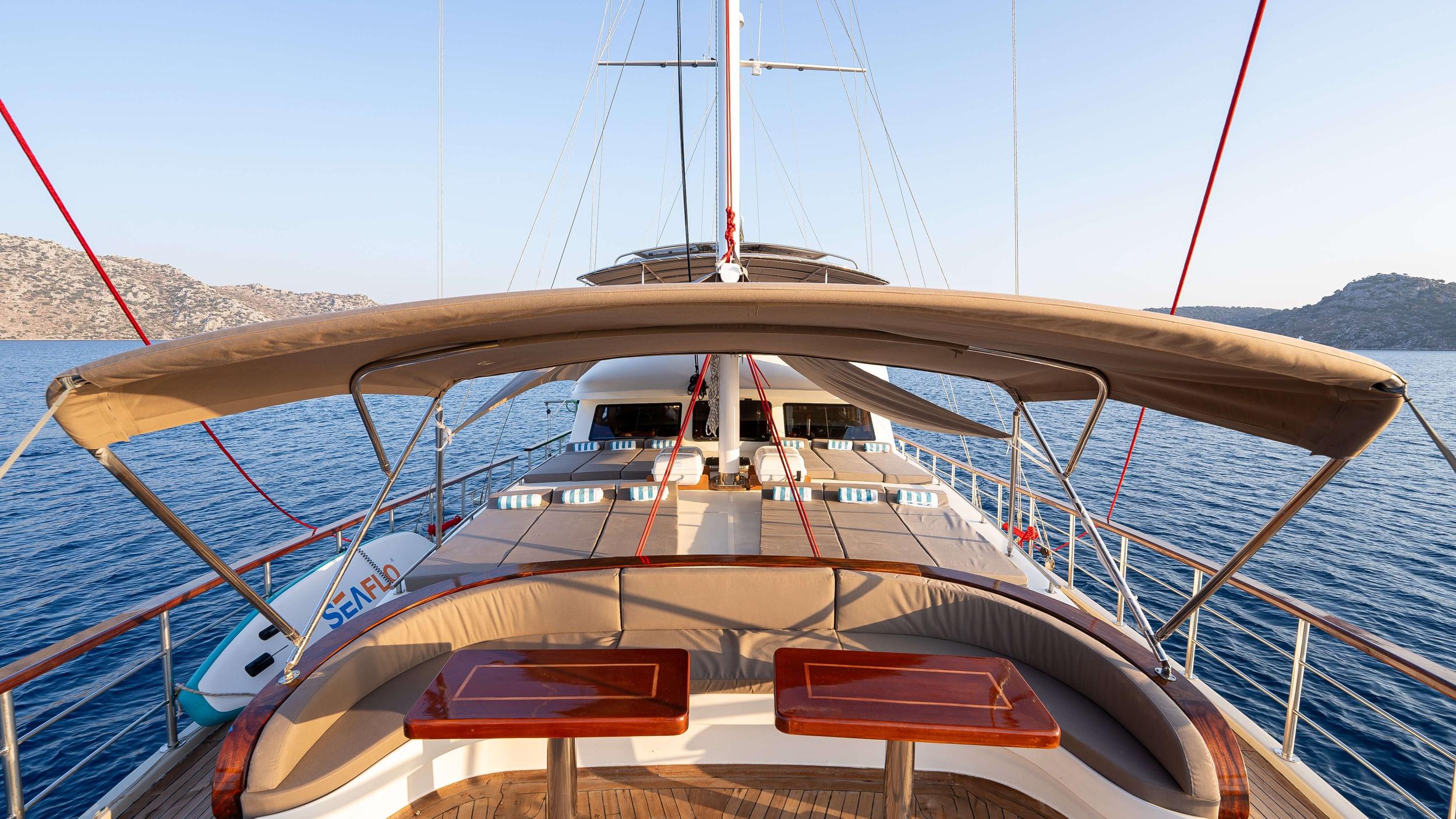 gulet bow deck featuring curved cushioned seating, teak decking, bimini awning, and paddleboard at anchor in calm bay