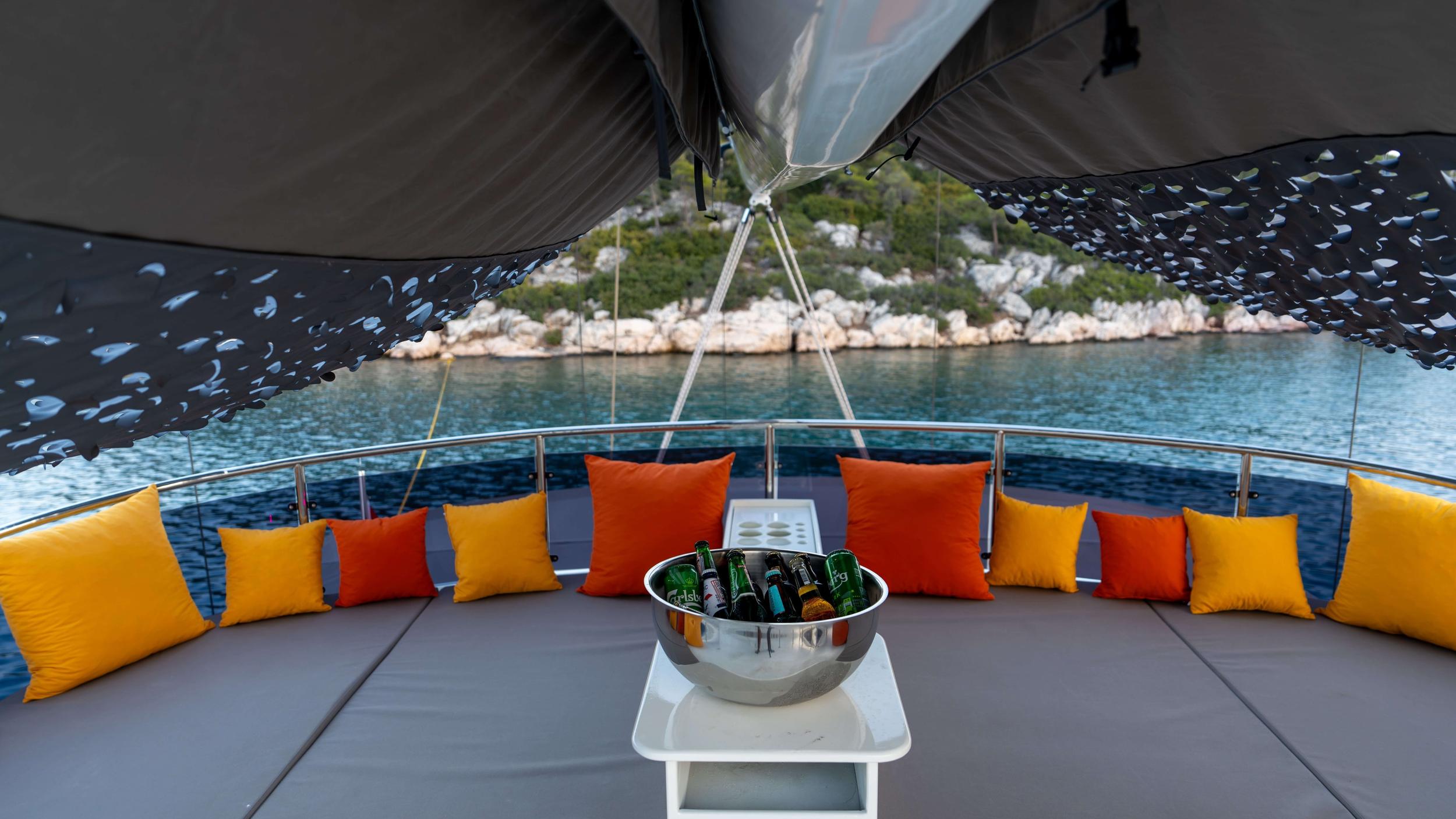 Gulet bow curved seating deck with orange and yellow cushions and chilled beer bottle bucket, anchored in rocky cove