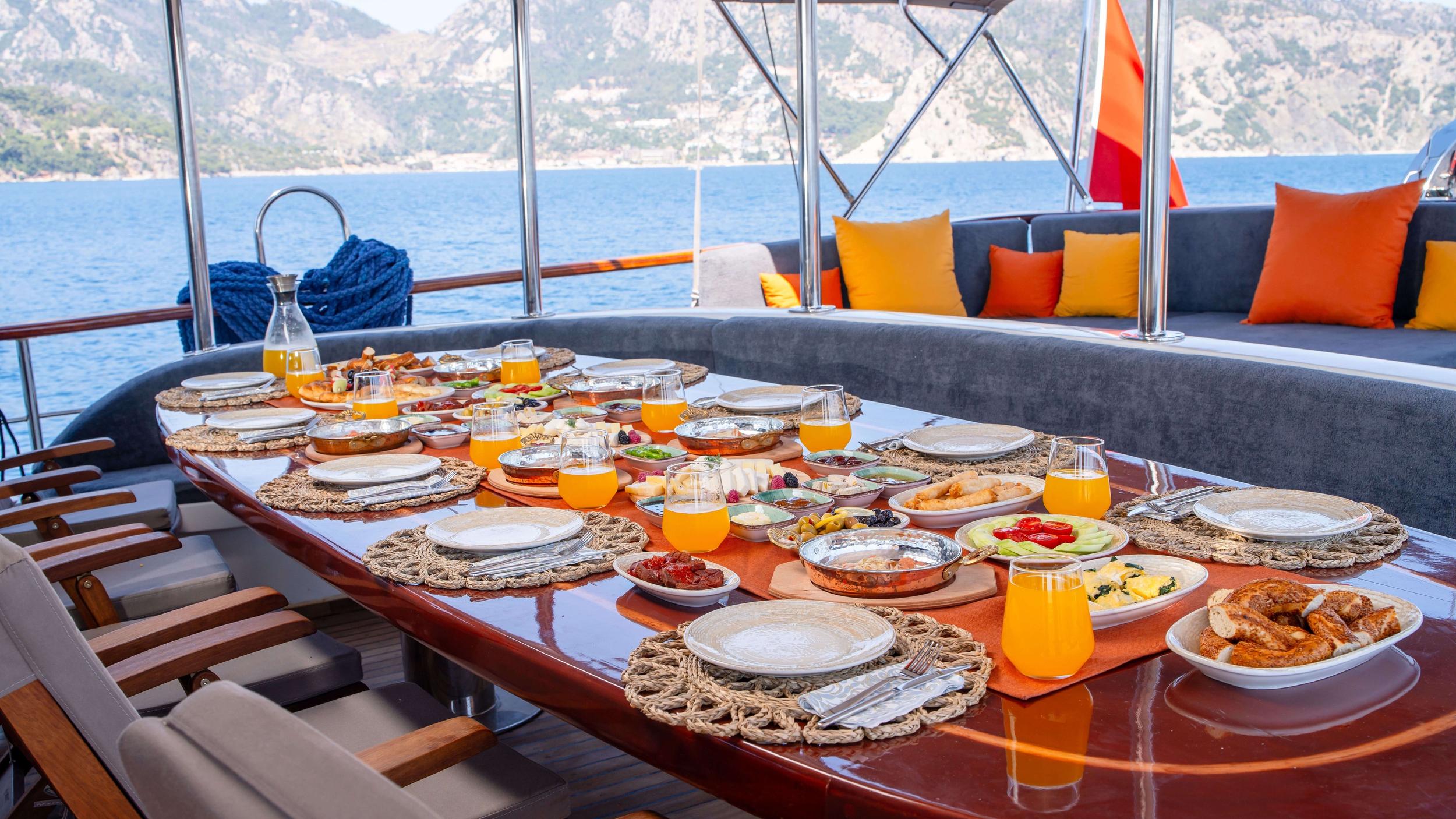 Luxury gulet aft deck dining table set with full Turkish breakfast spread, orange juice glasses, and wicker placemats with mountain backdrop