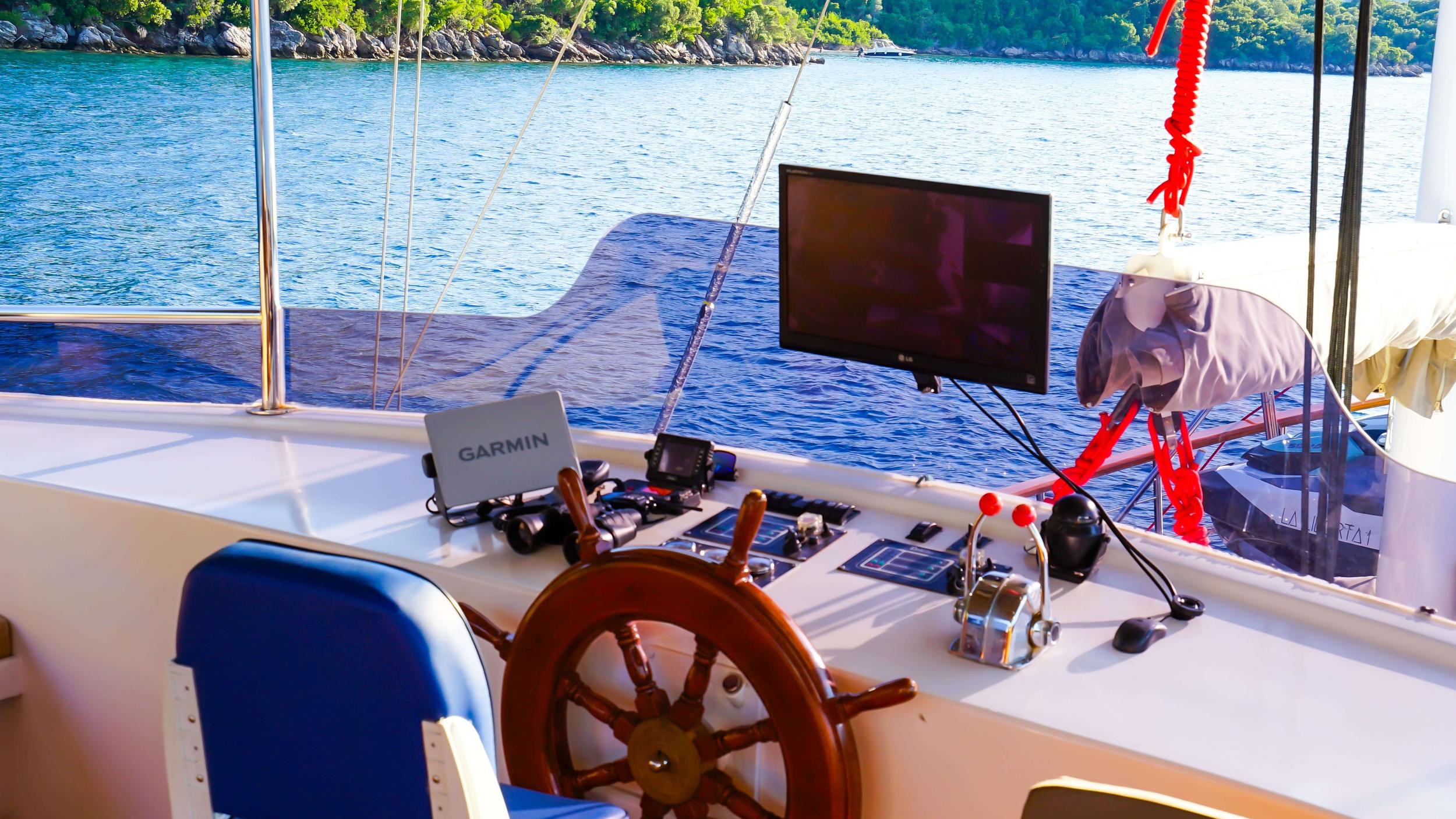 Gulet flybridge helm station with varnished wooden steering wheel, Garmin chartplotter, LG monitor, and throttle controls overlooking turquoise anchorage
