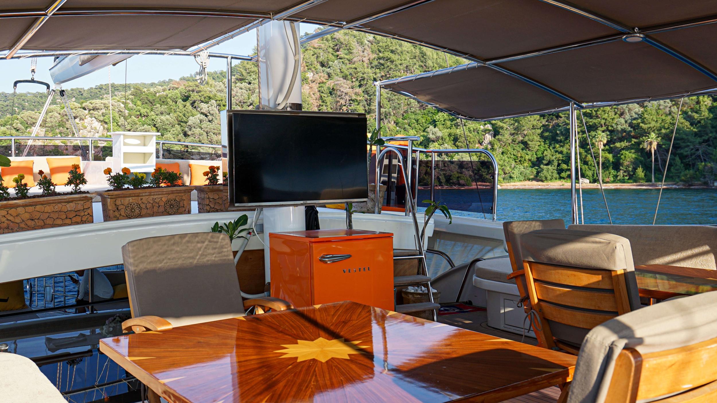 Gulet aft deck with varnished wood dining table, flat-screen TV mounted on mast, orange Vestel mini fridge, and canopy awning overlooking forested anchorage