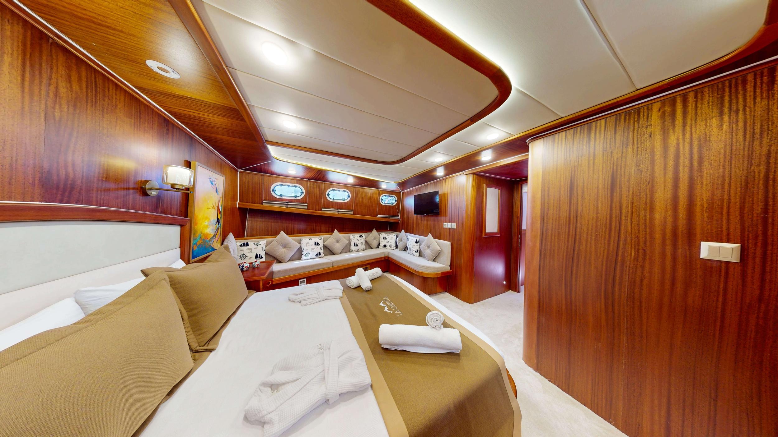 Spacious master cabin with double bed, L-shaped sofa seating area, oval portholes, flat-screen TV, and rich mahogany wood panelling