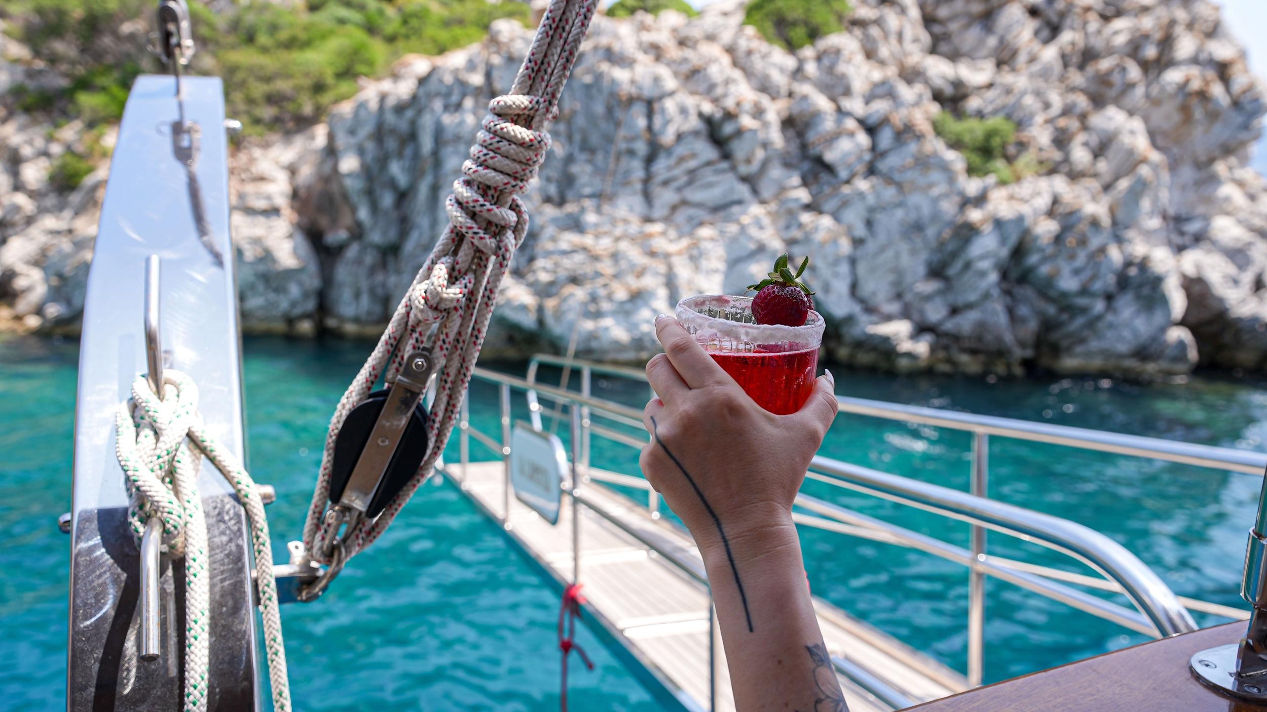 Hand holding a red cocktail garnished with a fresh strawberry on a gulet deck overlooking turquoise water and limestone cliffs