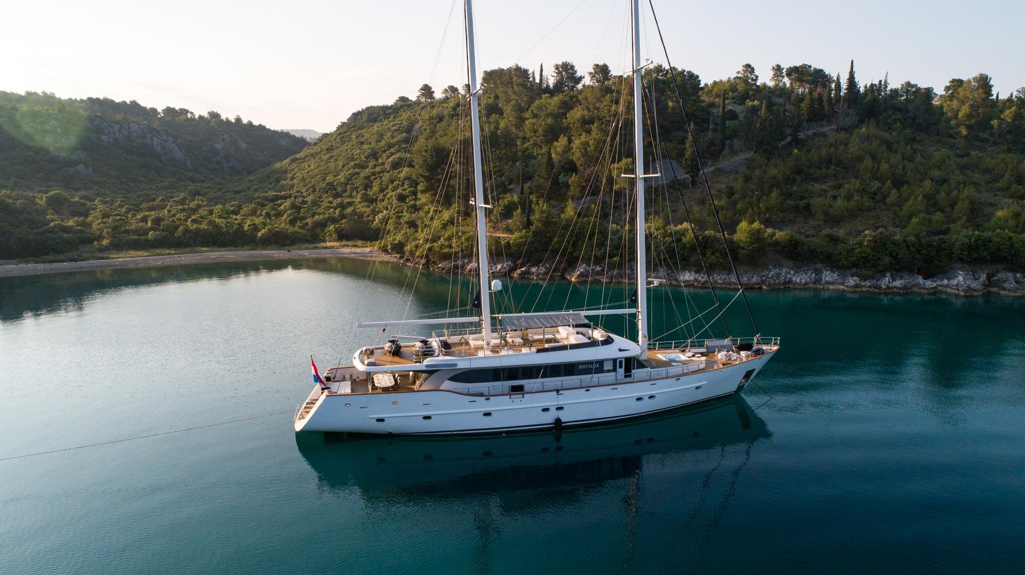 aerial view of white sailing yacht at anchor in turquoise Mediterranean bay surrounded by forested hills