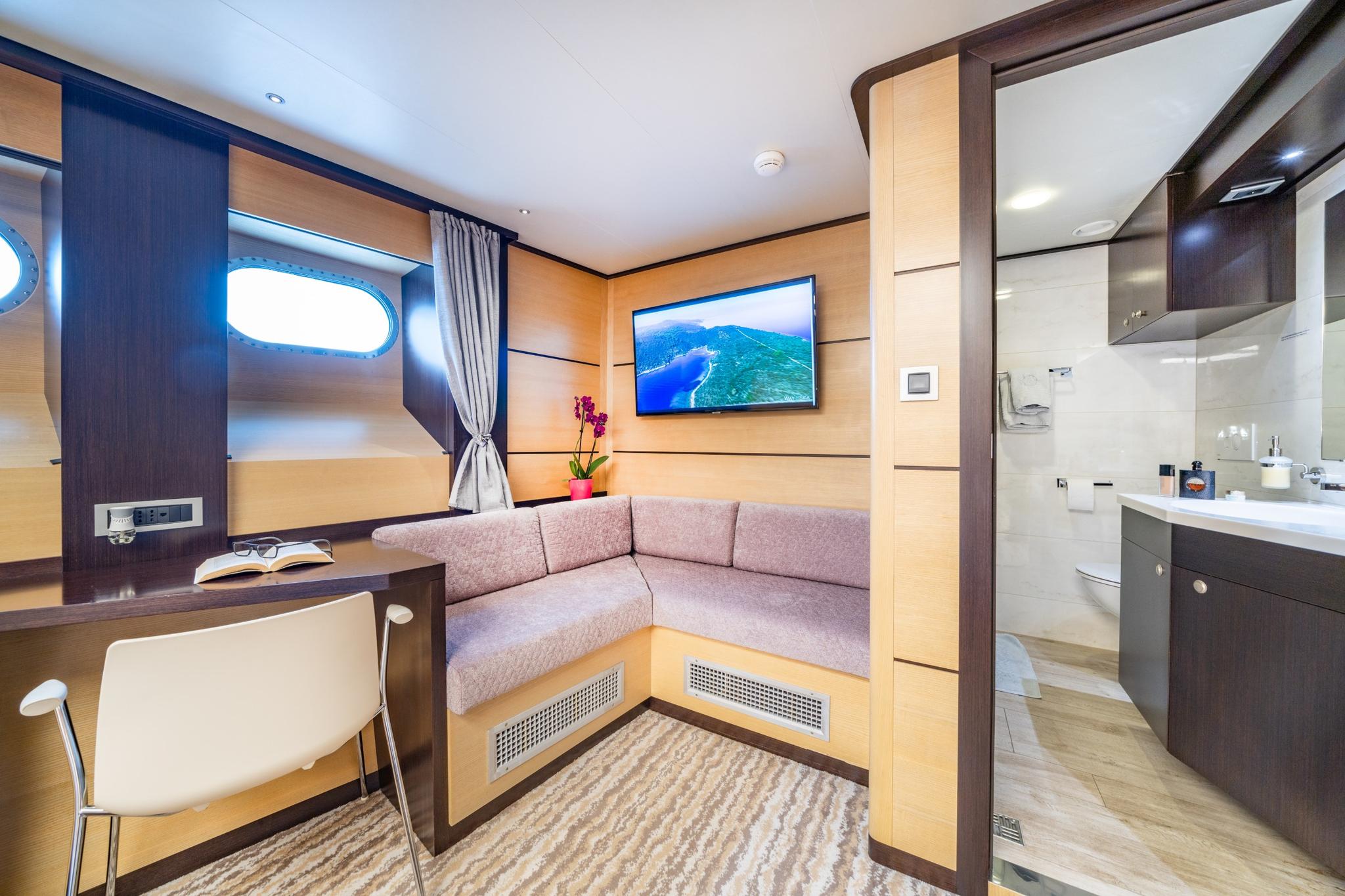 yacht cabin with corner seating, flat screen TV, oval porthole and ensuite bathroom visible