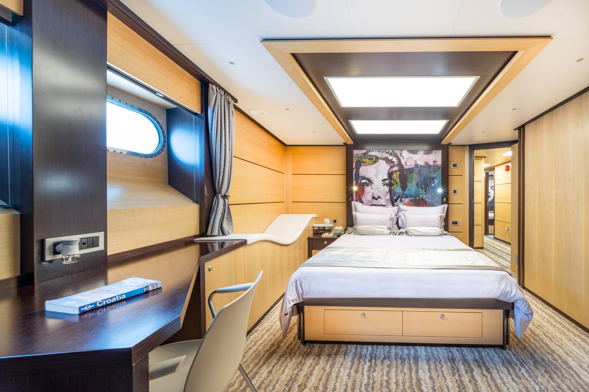 yacht cabin interior with double bed, colorful artwork headboard, work desk and porthole window