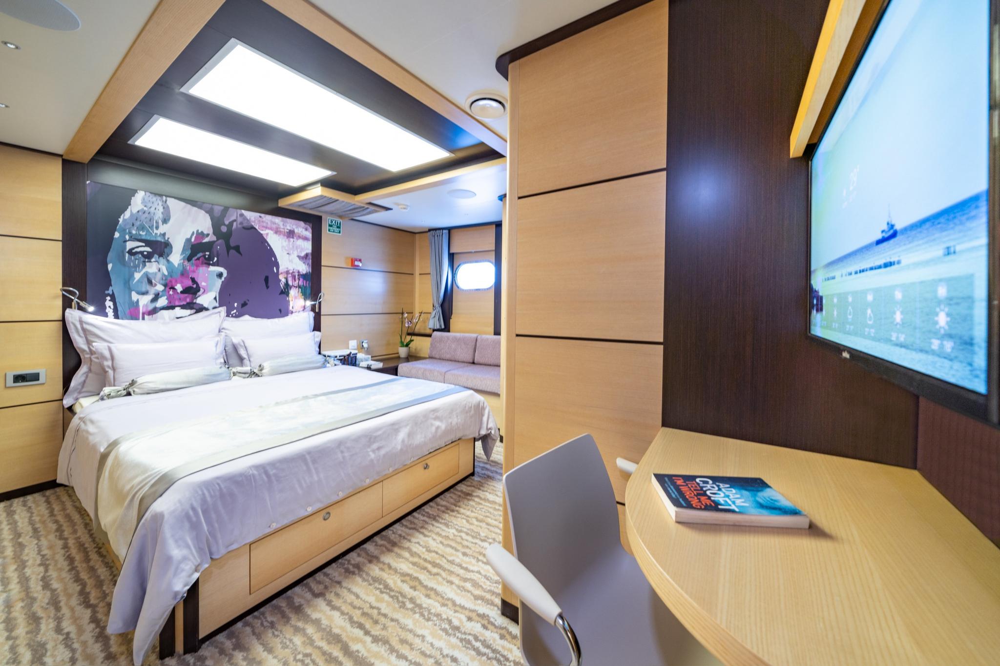 yacht cabin interior with double bed, colorful headboard artwork, wall-mounted TV and desk area