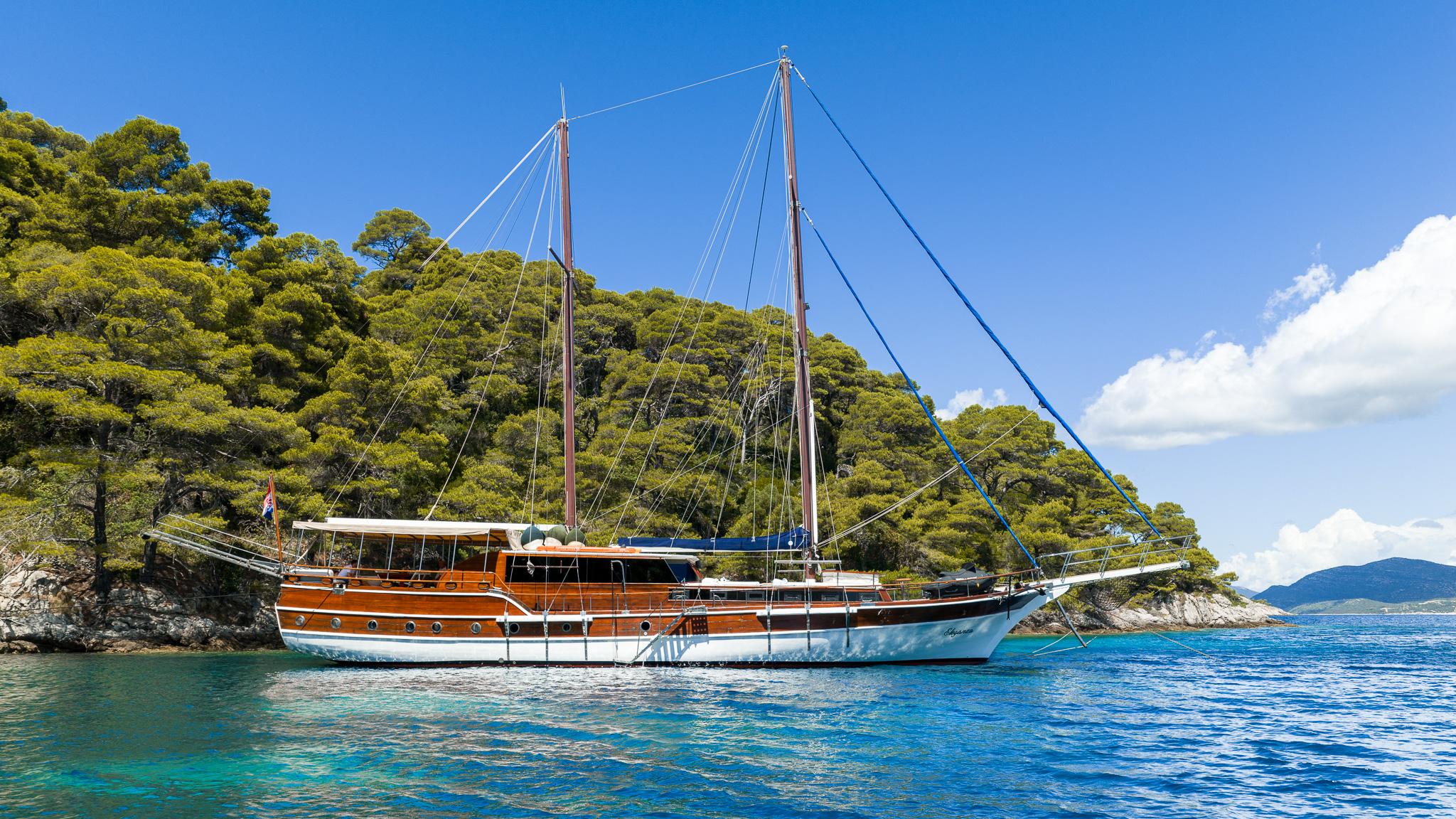 Traditional wooden gulet with two masts anchored in crystal clear turquoise waters near forested coastline