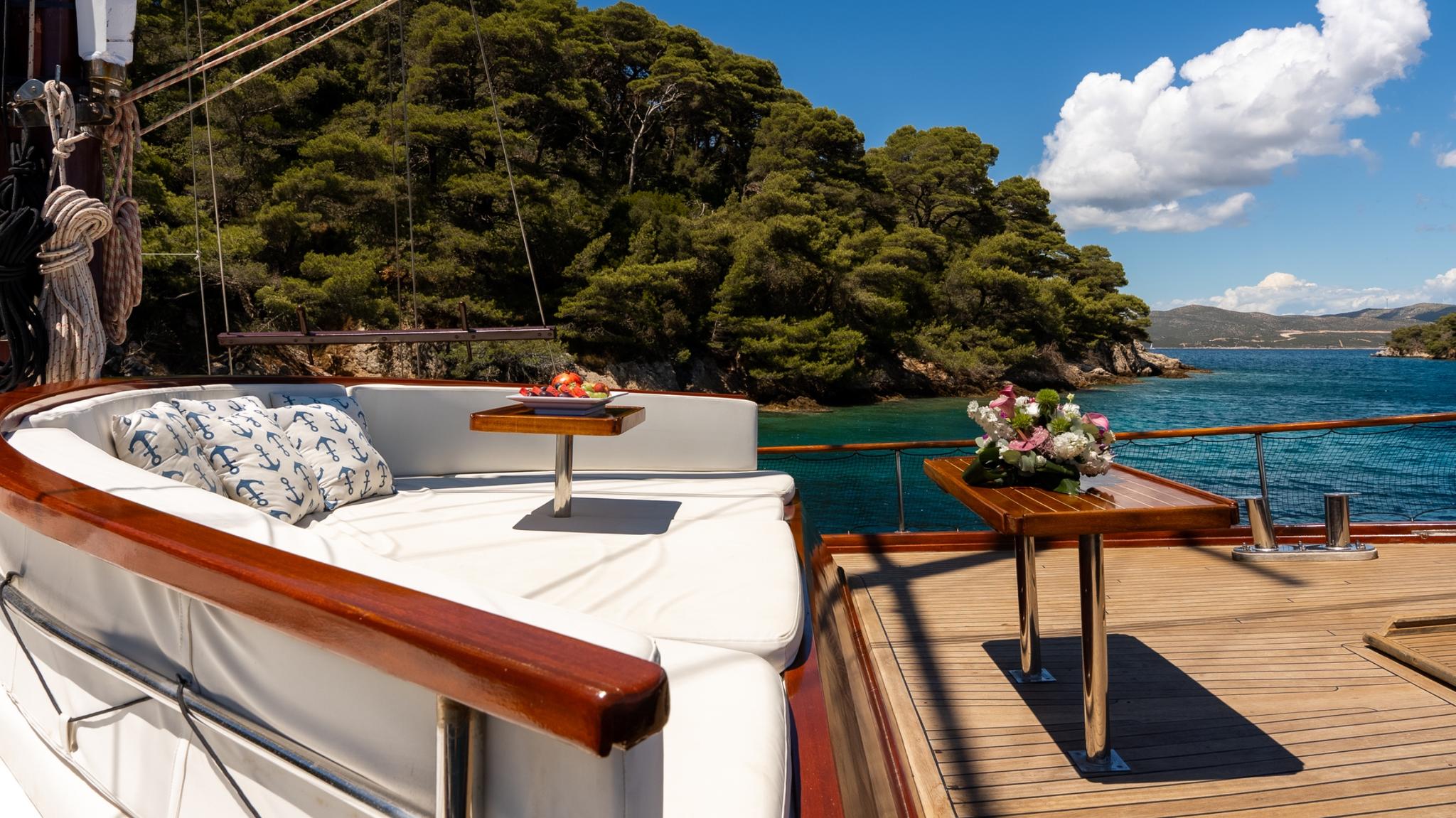 yacht deck seating area with white cushioned bench, wooden table, teak decking overlooking turquoise Mediterranean coastline