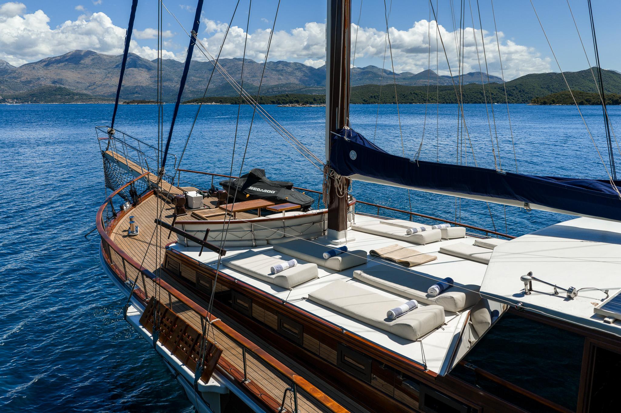 gulet sun deck featuring white cushioned loungers arranged around mast with rigging and mountainous coastline