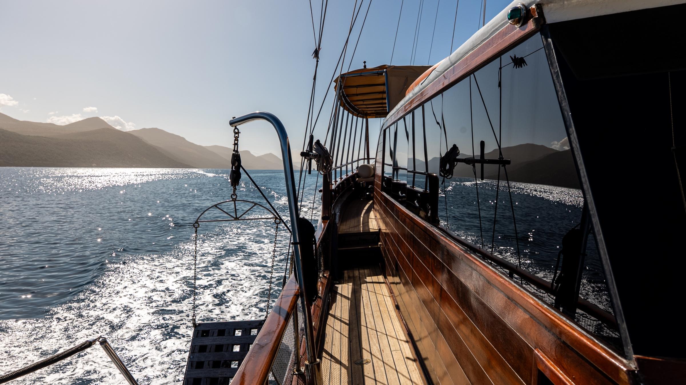 Traditional wooden gulet deck with rigging and railings sailing past mountainous coastline