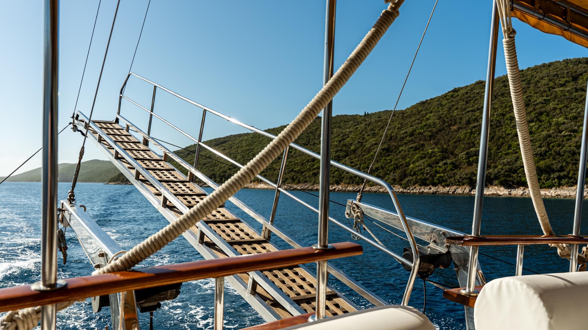 bow deck of sailing yacht with wooden rigging, stainless steel railings and Mediterranean coastline