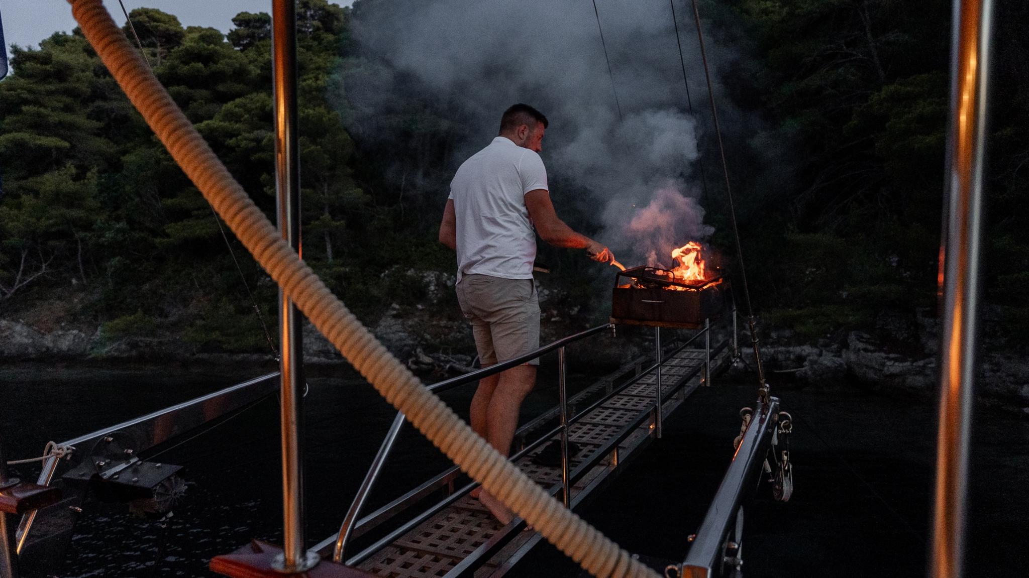 crew member grilling food on barbecue at yacht stern with flames visible