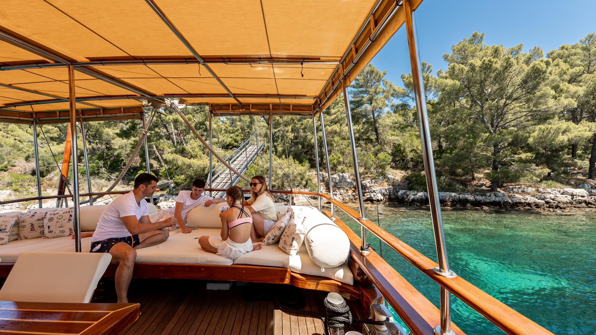 Gulet aft deck with cushioned seating area, canvas awning, guests relaxing in Mediterranean cove