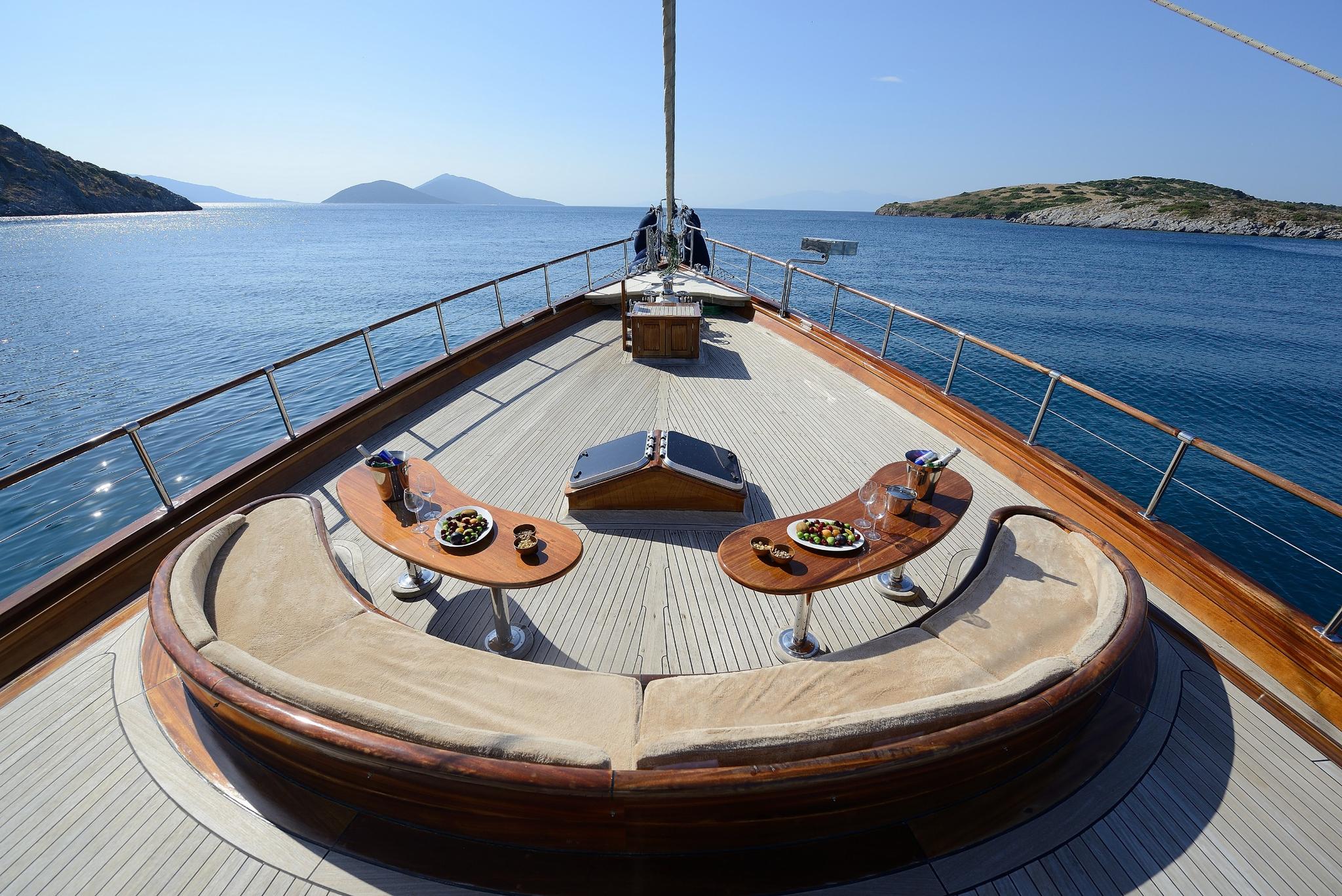yacht bow deck with curved built-in seating, wooden tables, and Mediterranean coastline views