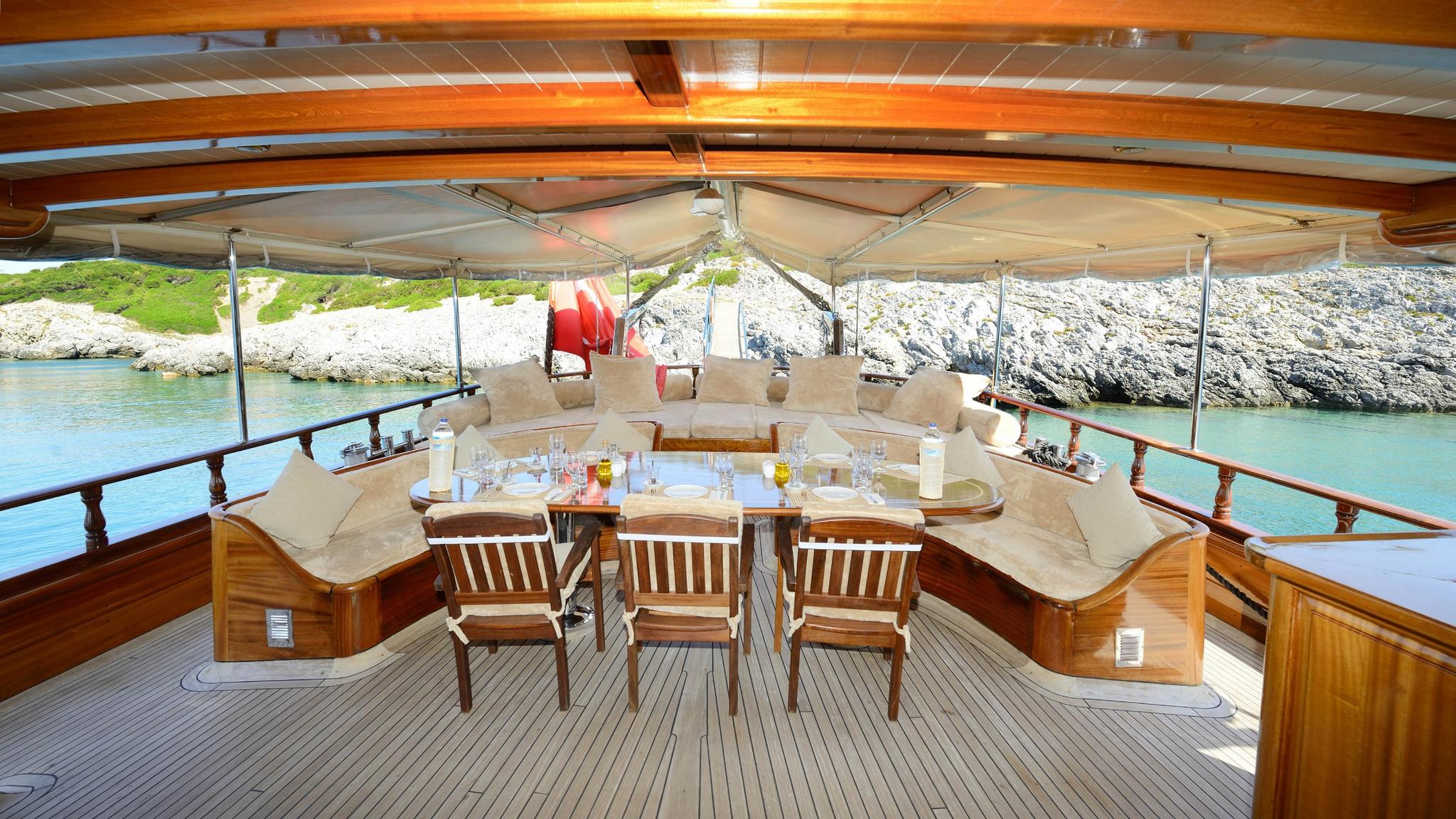 Gulet aft deck dining area with curved seating, wooden table and coastal backdrop