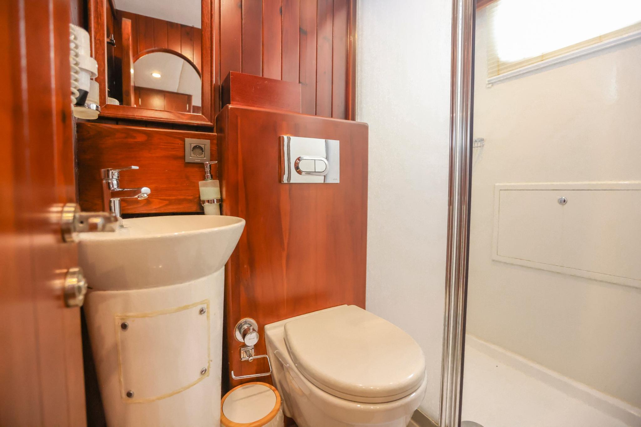 Yacht bathroom with white ceramic sink, toilet, glass shower door and mahogany wood paneling