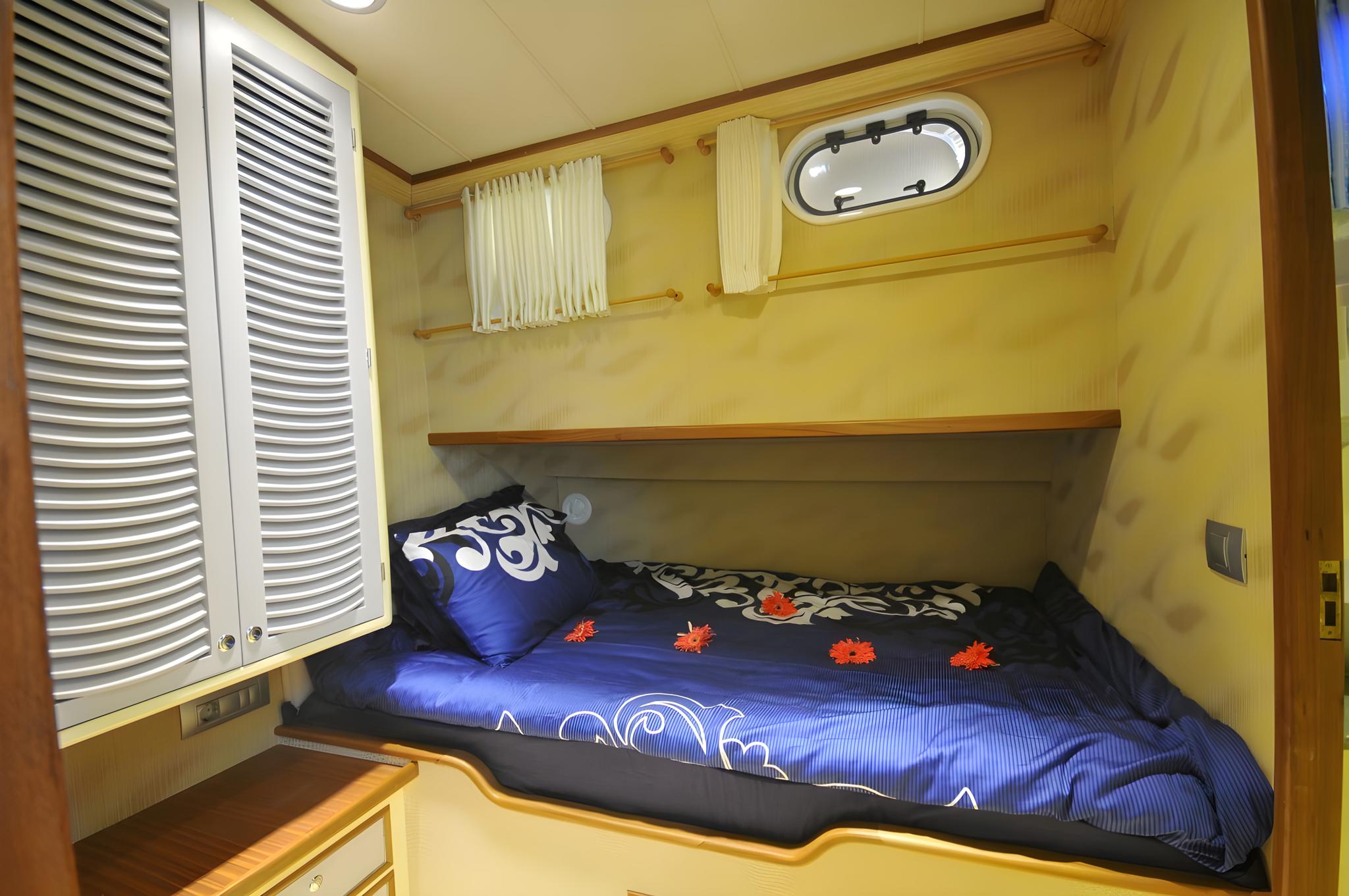 yacht guest cabin featuring double bed with blue bedding, oval porthole window, and white louvered storage doors