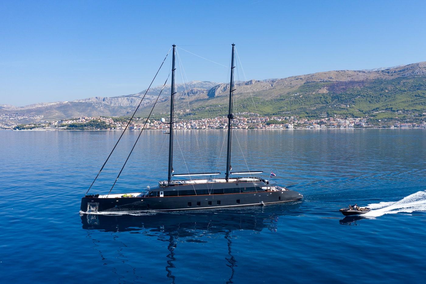 Large sailing yacht with two masts anchored in blue waters near mountainous coastline