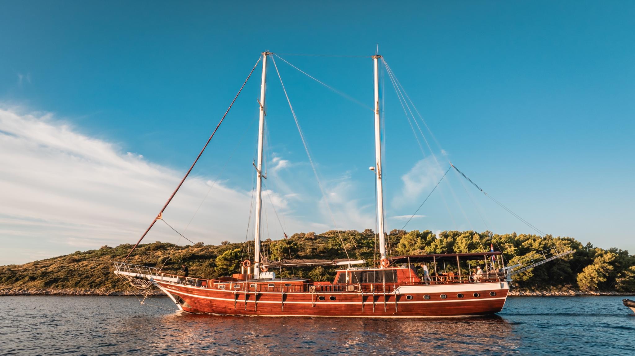 Traditional wooden gulet yacht with reddish hull anchored in calm waters near forested coastline