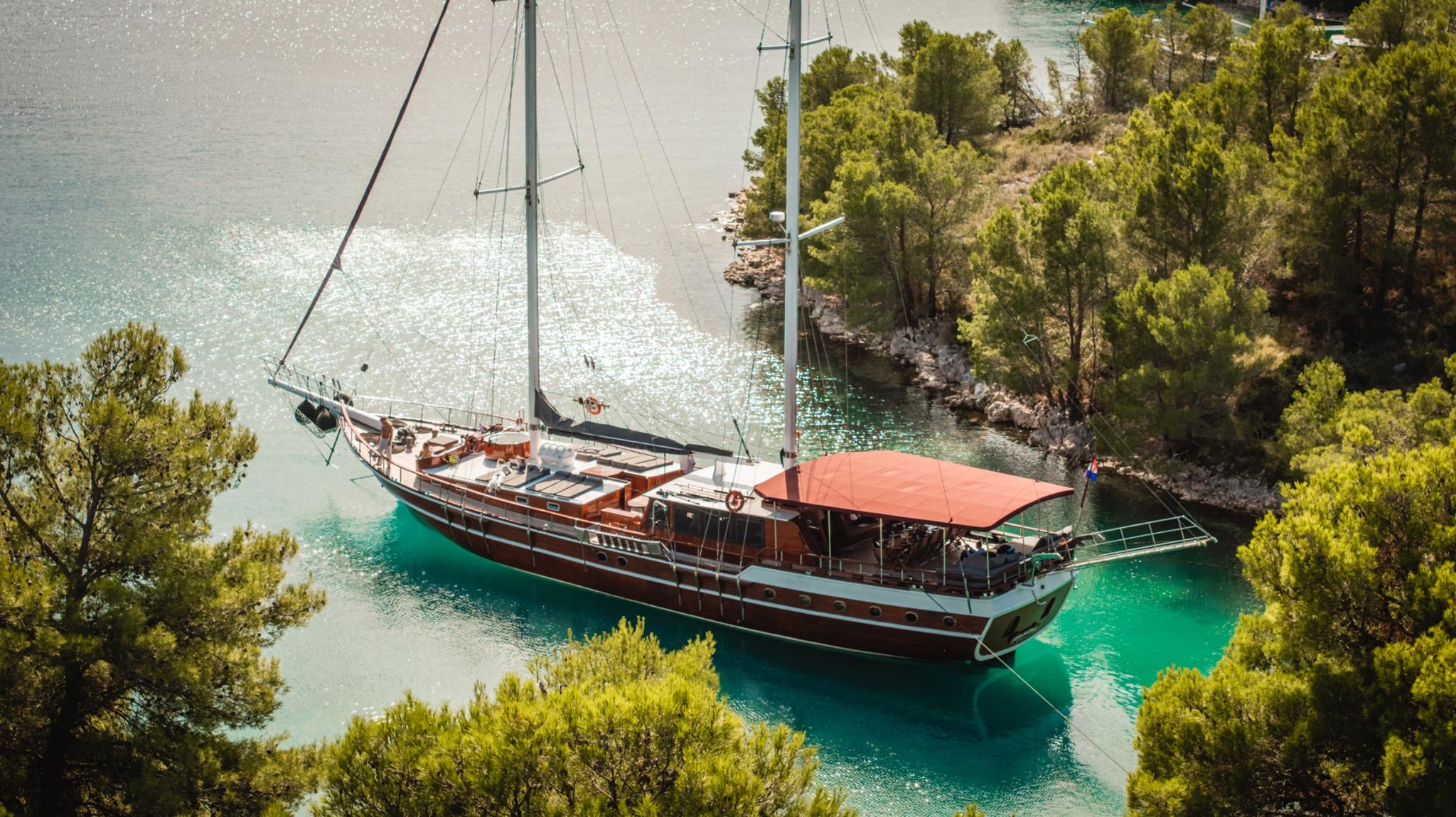 aerial view of wooden gulet with red awning anchored in crystal clear turquoise bay surrounded by pine trees