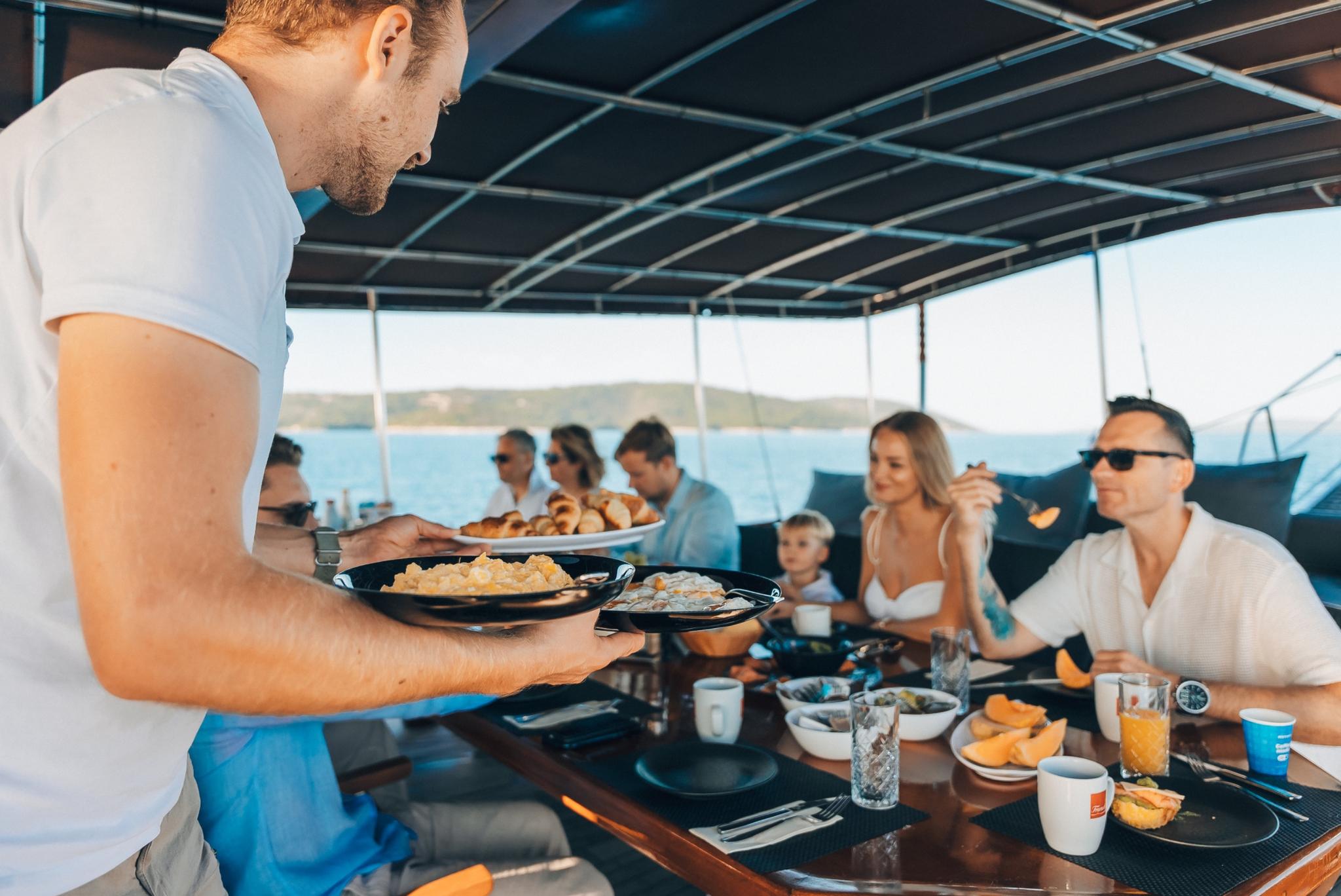 crew member serving cooked breakfast from black plates to guests dining on covered deck area