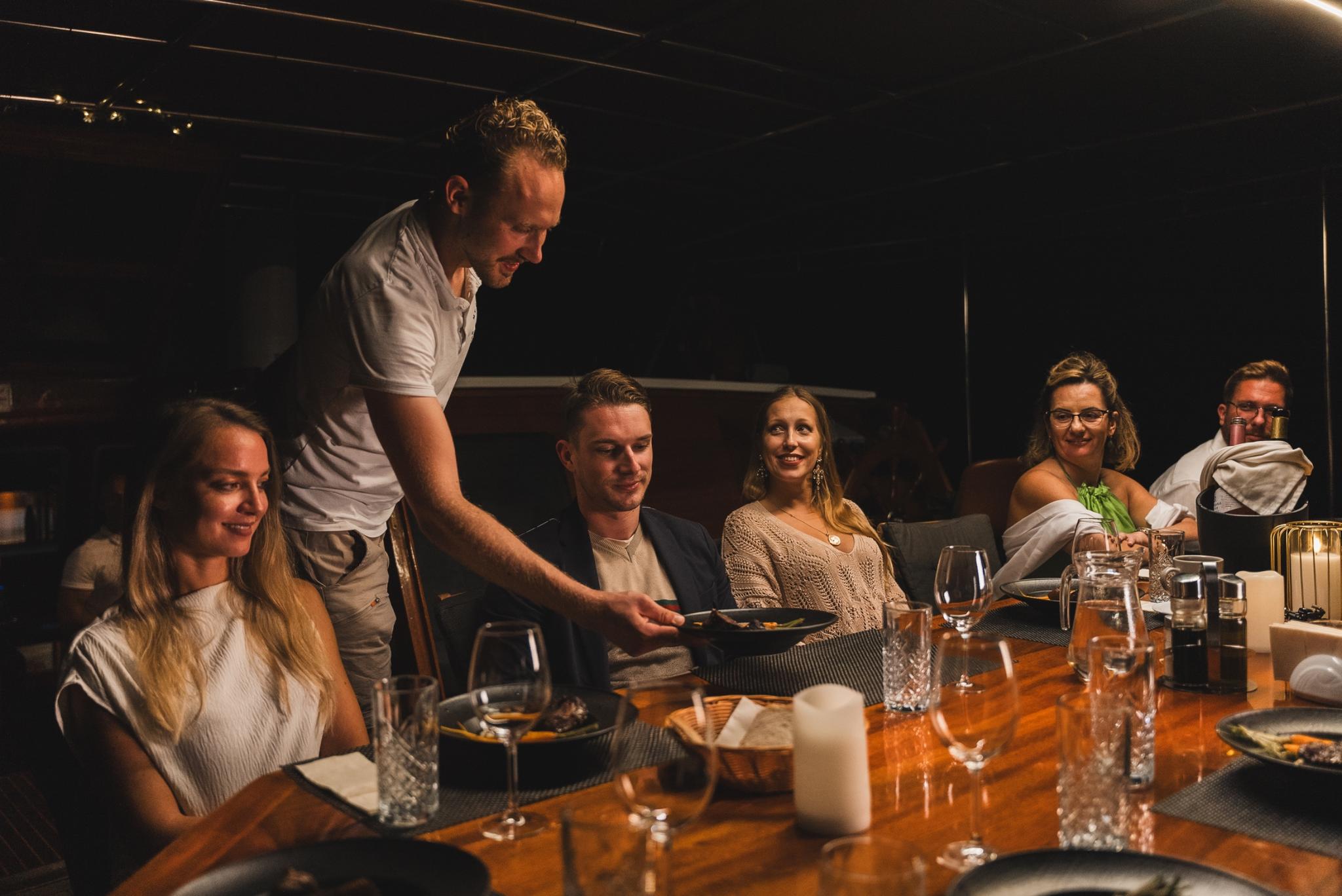 crew member in white polo shirt serving plated dinner to guests seated around yacht dining table
