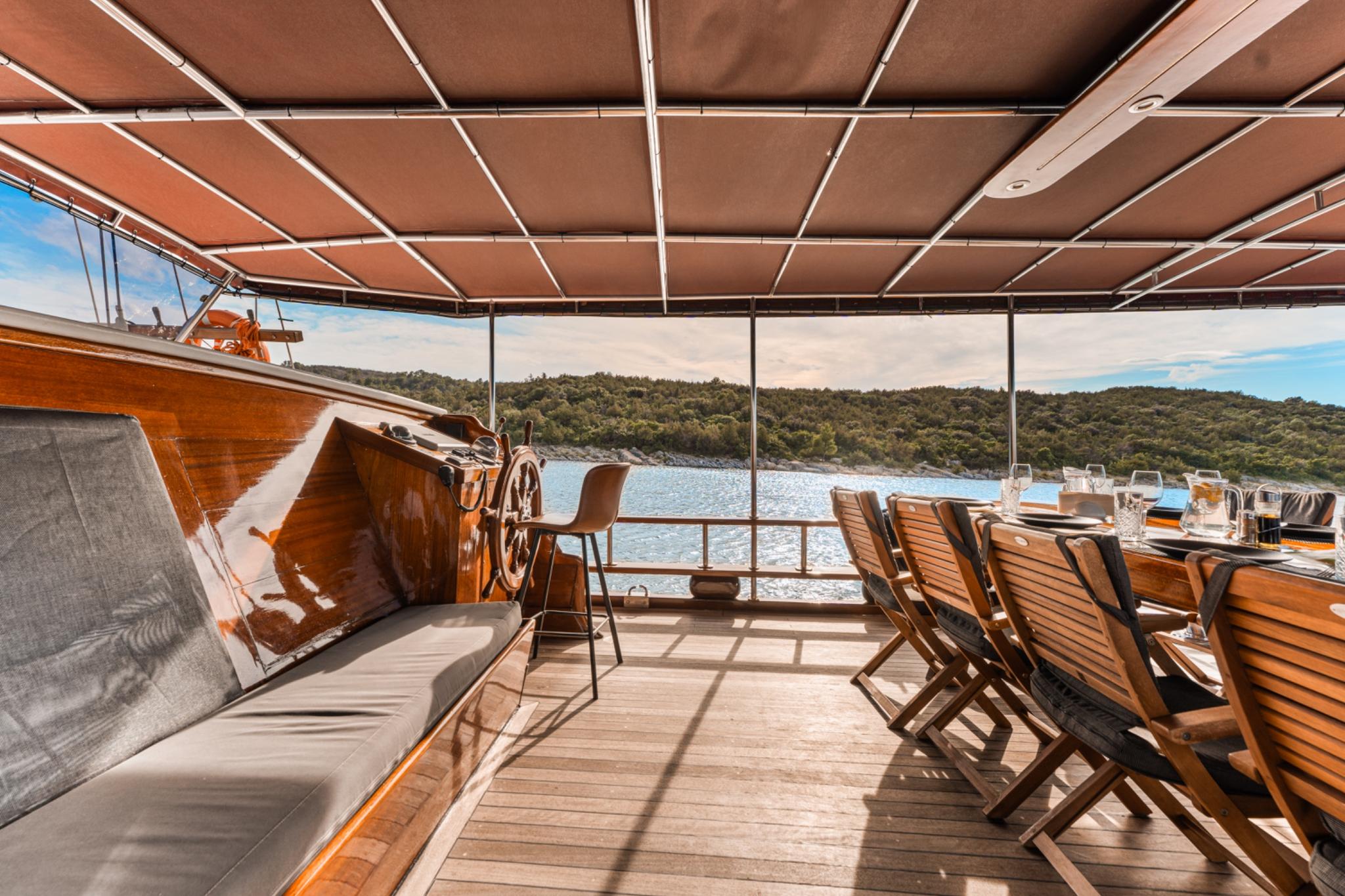 luxury gulet aft deck with wooden dining chairs, canvas awning, and helm station overlooking forested coastline