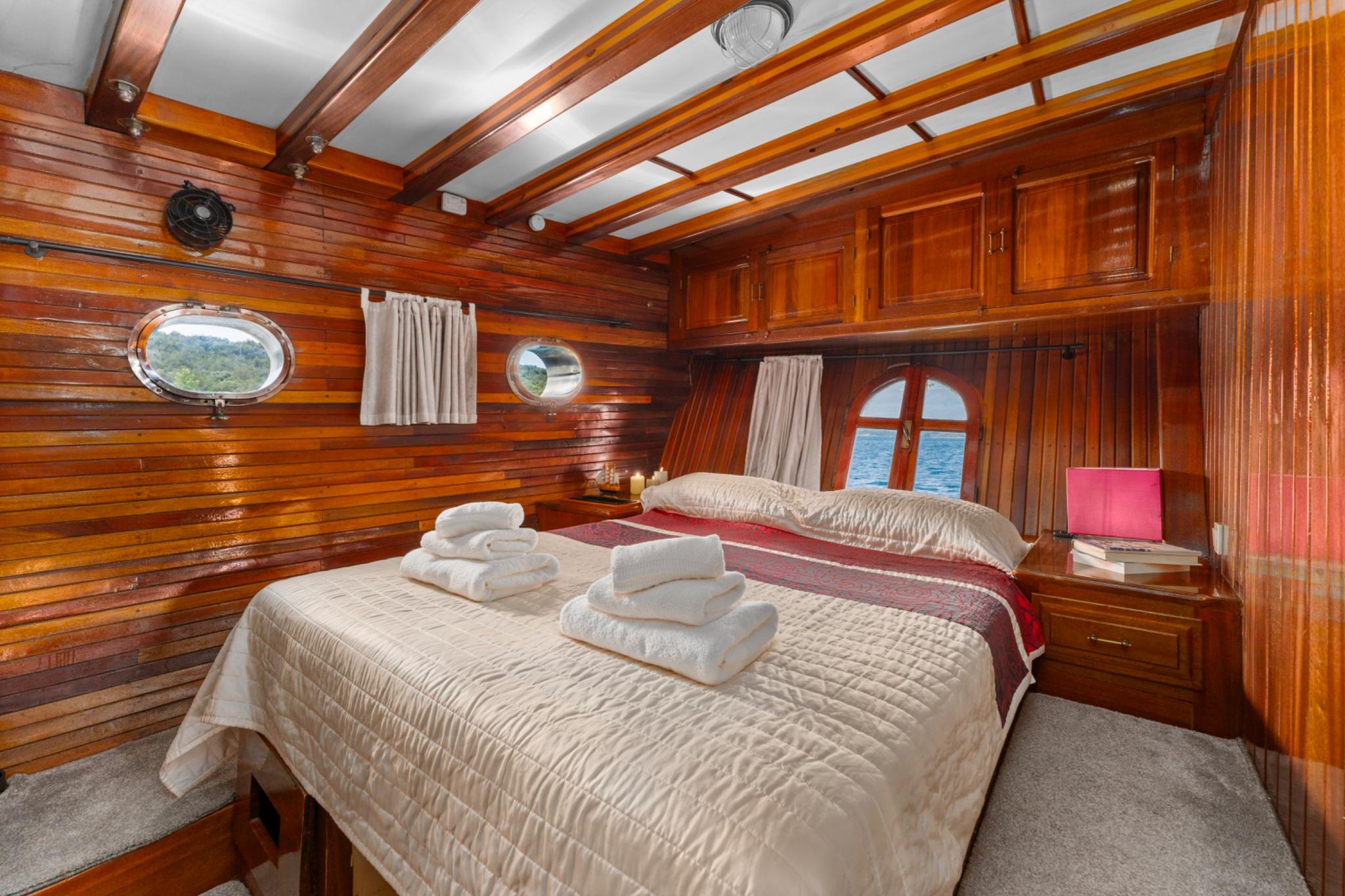 yacht cabin interior with double bed, white linens, towels, wooden paneling and circular portholes