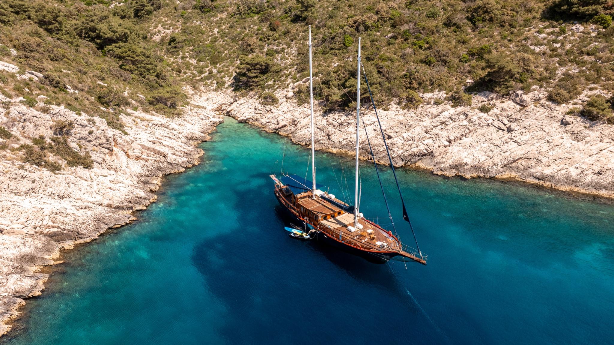 Aerial view of wooden gulet yacht anchored in turquoise Mediterranean cove with limestone cliffs