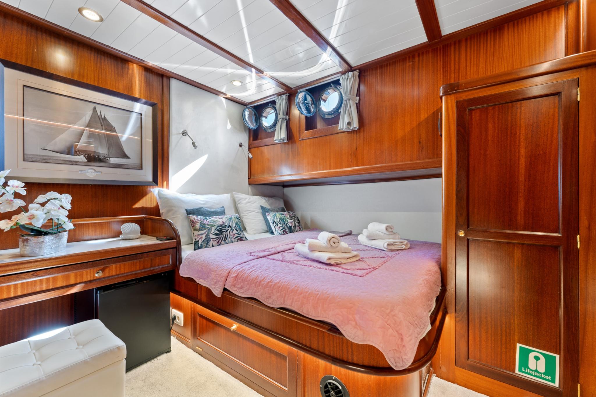 yacht cabin interior featuring double bed with pink bedding, rich teak wood paneling, two portholes, and built-in wardrobe