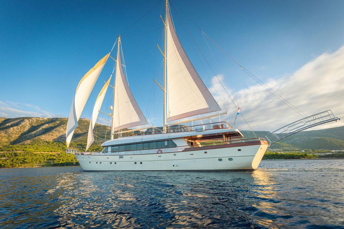 White luxury gulet with sails up anchored in calm bay with forested hills