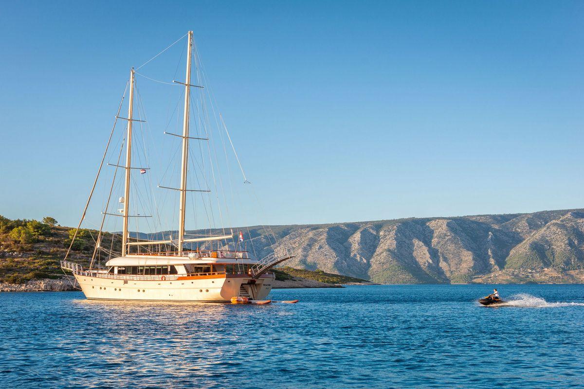 Traditional wooden gulet yacht anchored near rocky coastline with tender boat