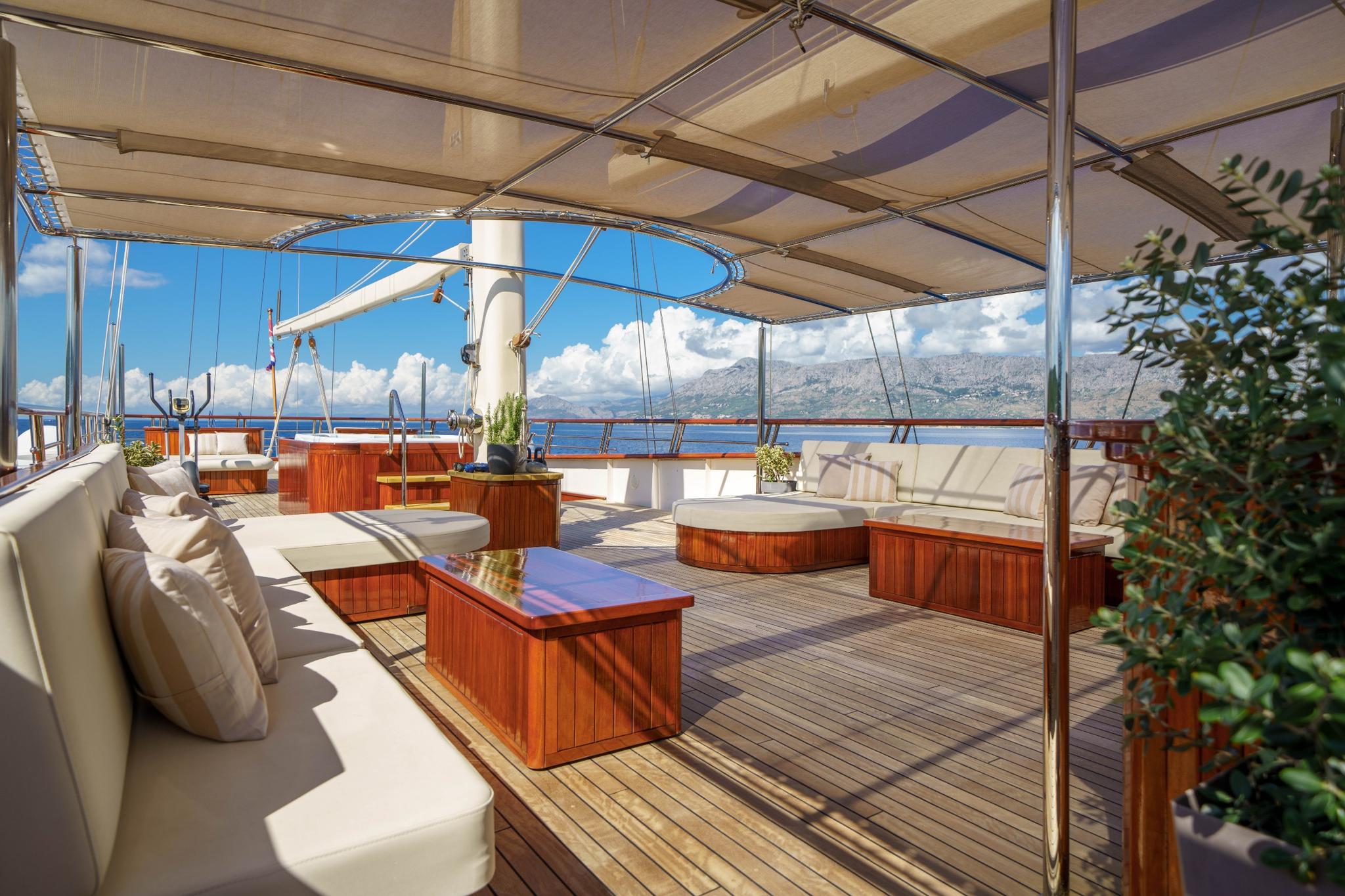 gulet upper deck lounge with white cushioned seating, teak coffee tables and mountain coastline views
