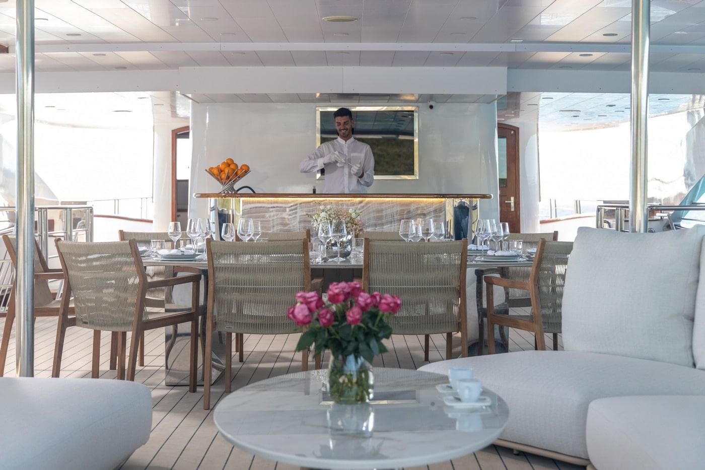 Luxury yacht covered dining area with formal table setting and crew member