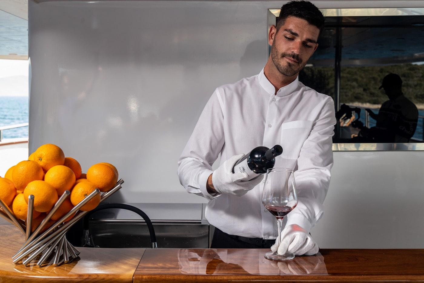 yacht crew member in white uniform pouring red wine glass onboard