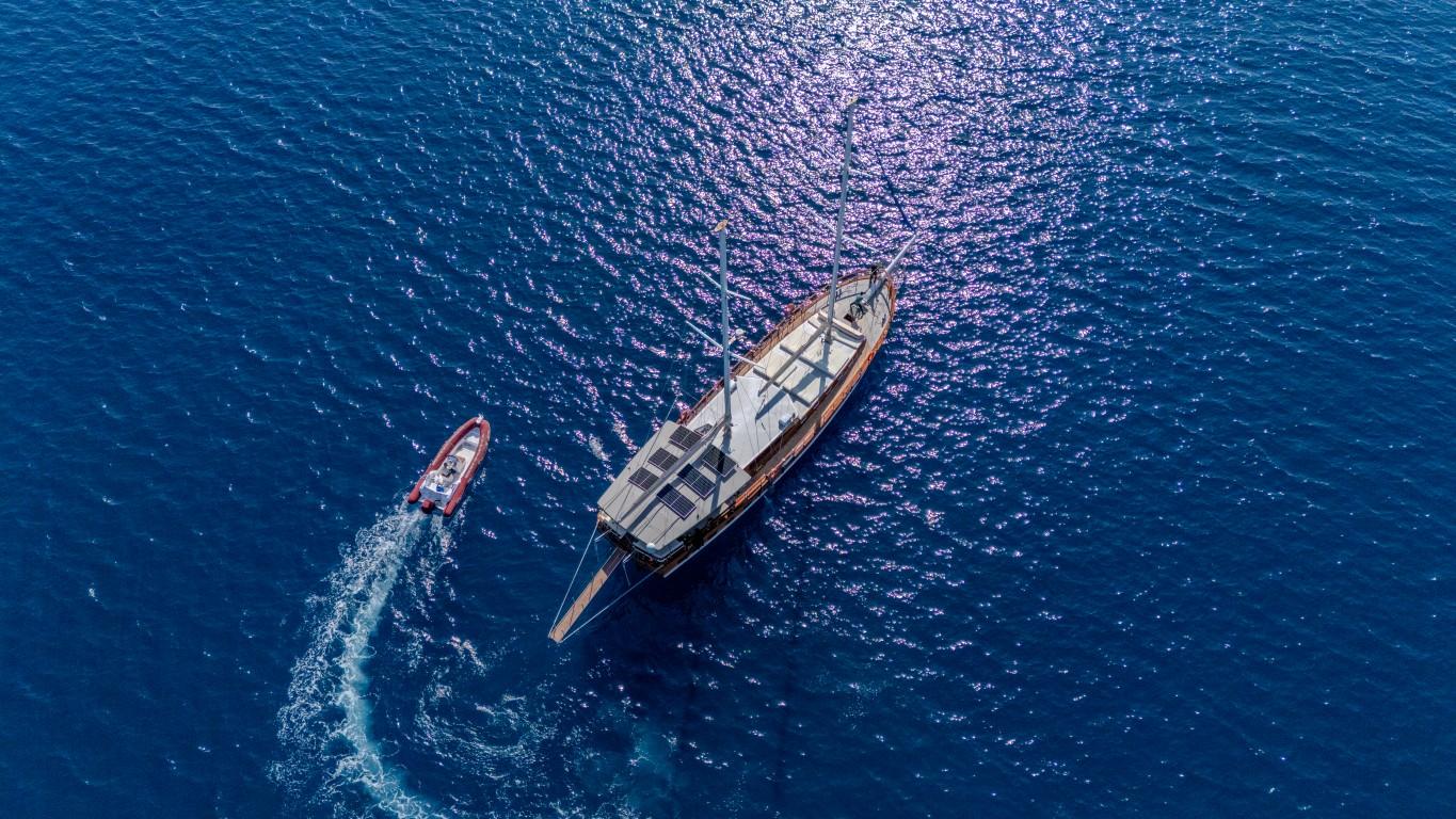 aerial view of traditional gulet yacht at anchor with red tender boat creating white wake trail