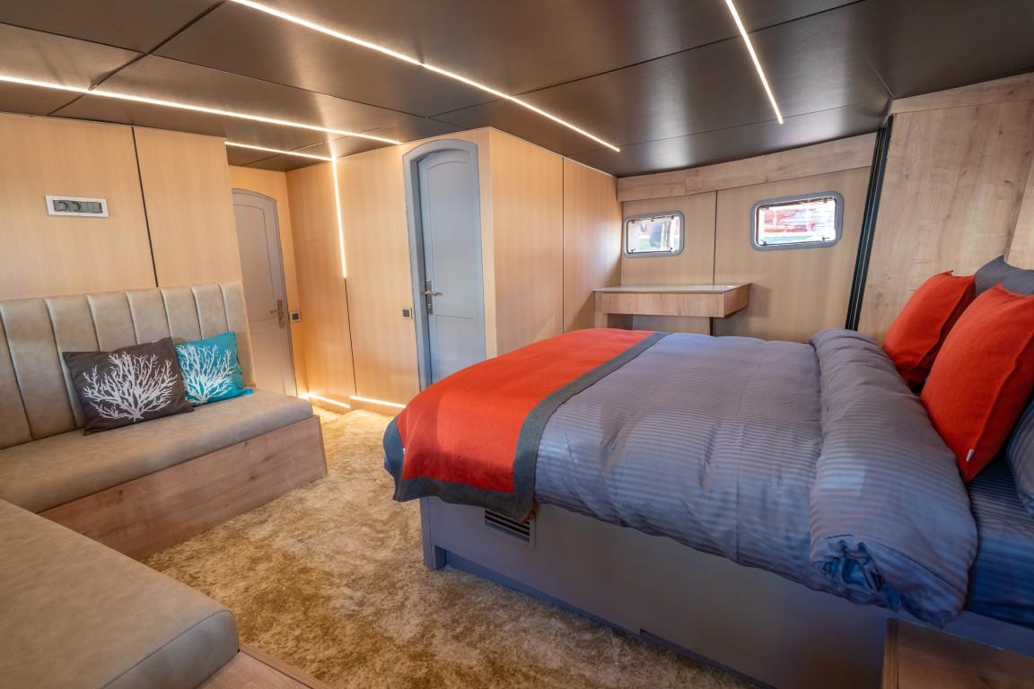 Spacious yacht cabin featuring double bed with red and grey bedding, built-in seating with coral cushions