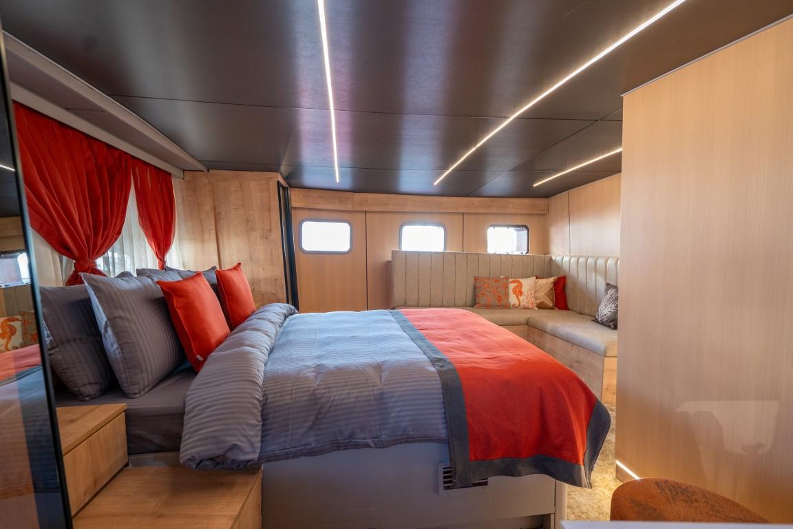 yacht cabin interior featuring double bed with red and grey bedding, built-in seating area with cushions, and three portholes