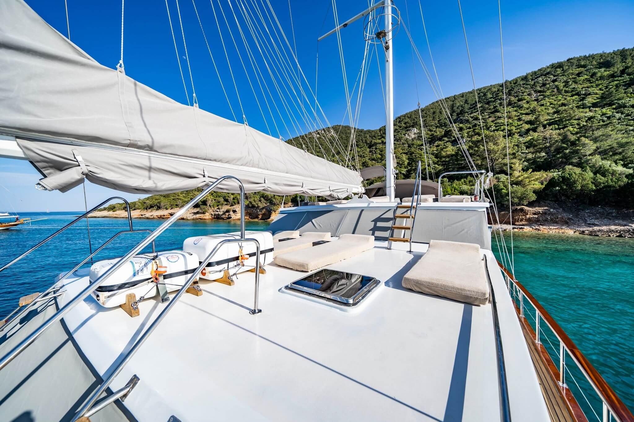 Gulet bow deck featuring cream cushioned sun loungers, furled mainsail, white mast rigging, and a flush deck hatch, anchored in a turquoise cove with pine-covered hillside