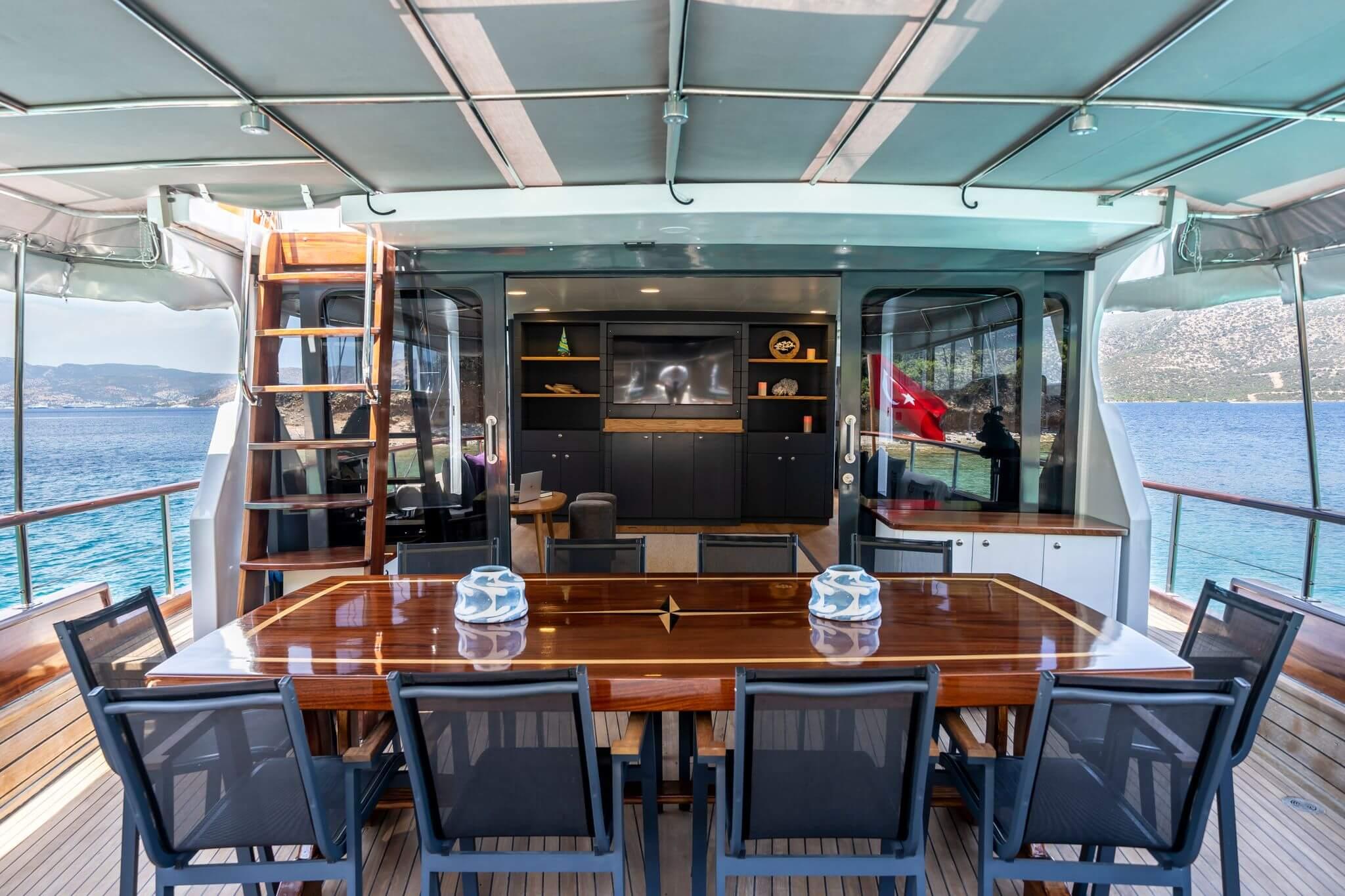 Gulet aft deck with polished teak dining table seating eight chairs under grey canopy with black cabinetry and flatscreen TV visible through sliding doors