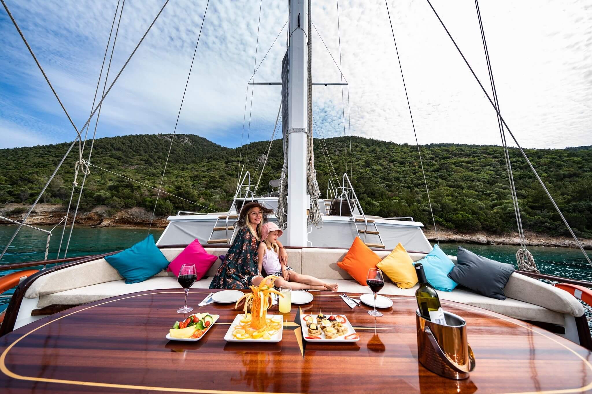 woman and child seated on cushioned foredeck sofa beside polished teak table laid with mezze plates and red wine glasses