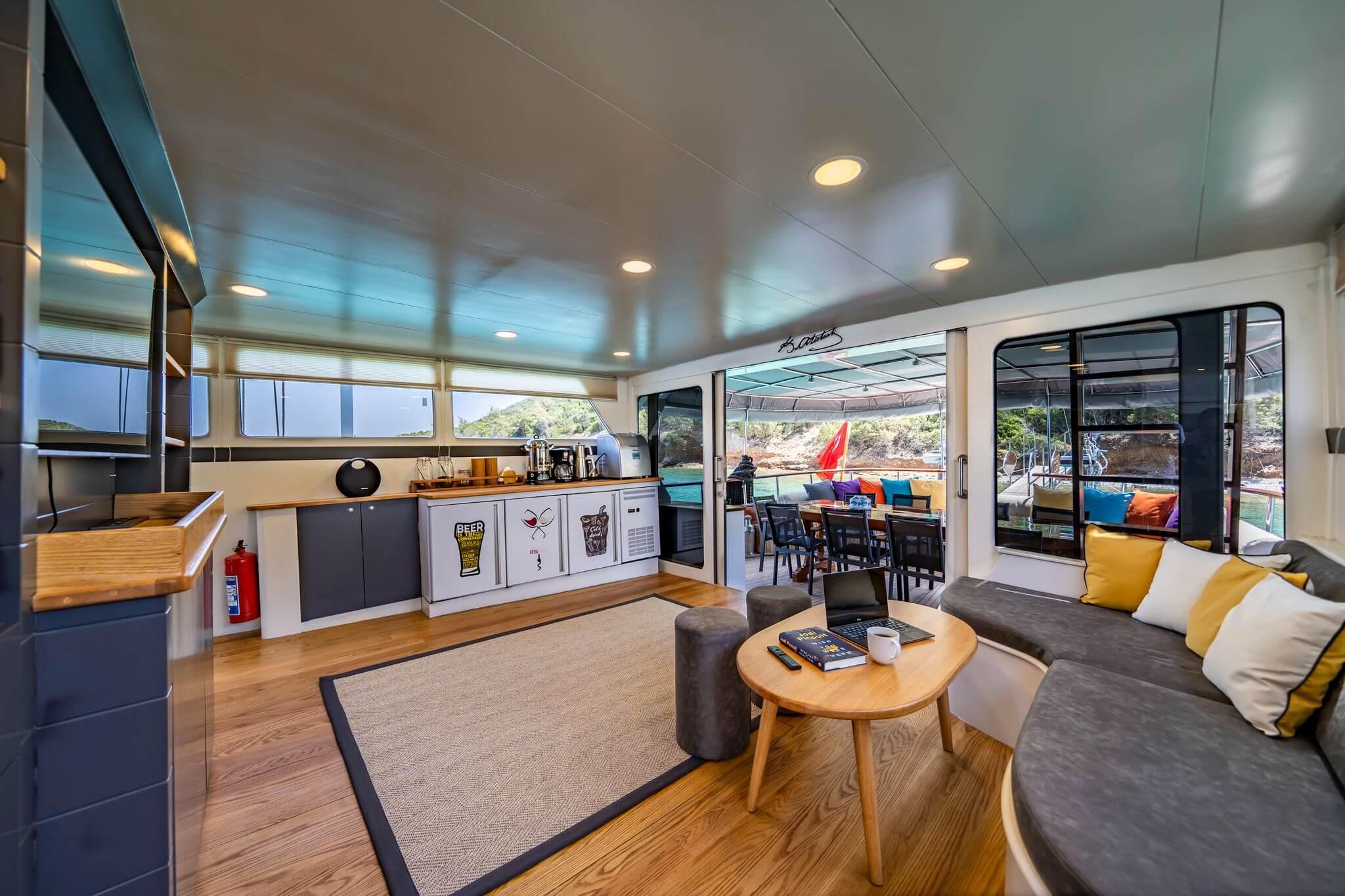Gulet indoor salon with curved grey sofa, round coffee table, and galley bar counter opening to aft deck dining area