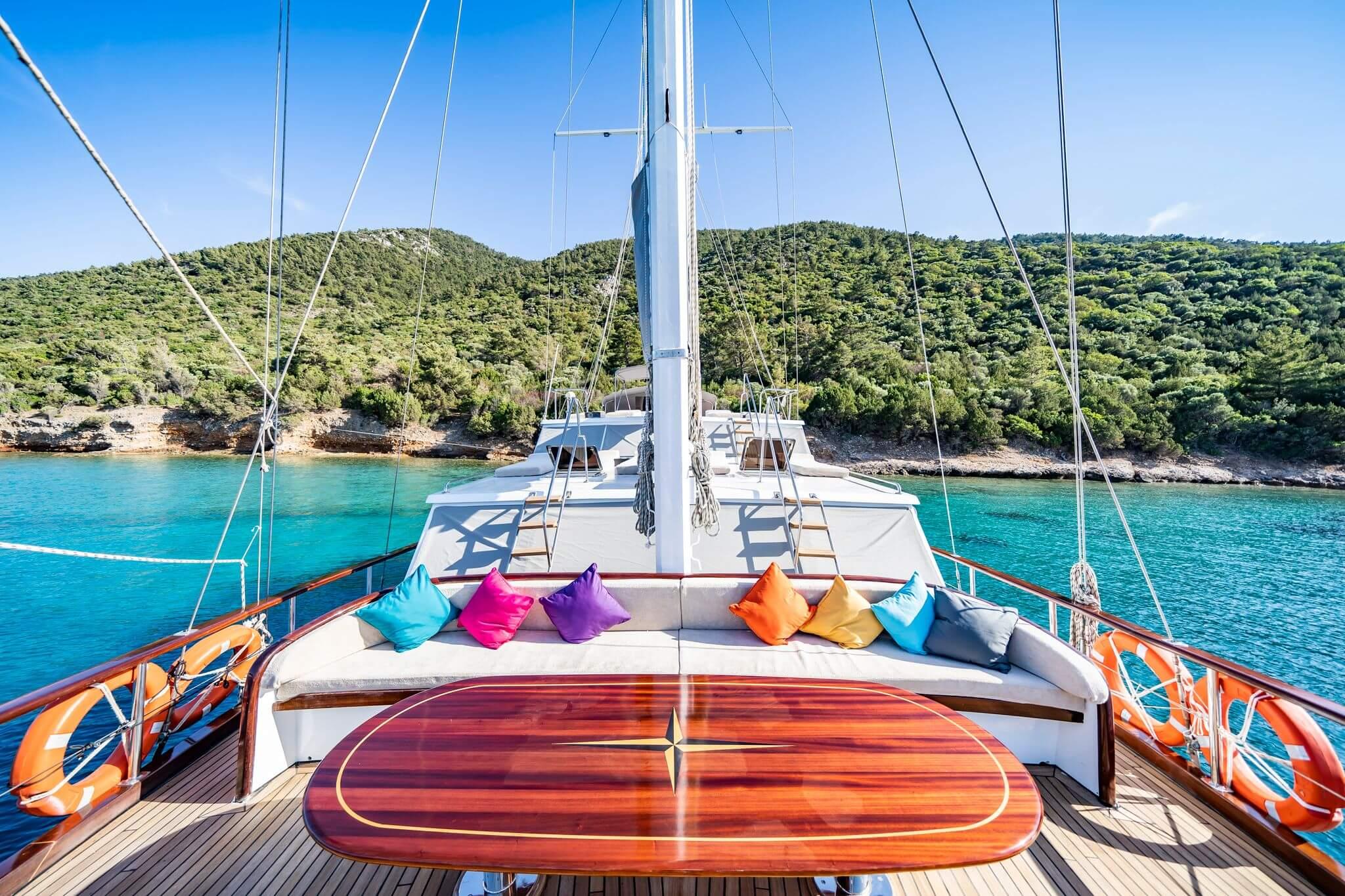 Gulet bow deck with colourful scatter cushions, polished teak compass-rose table, and turquoise anchorage beyond
