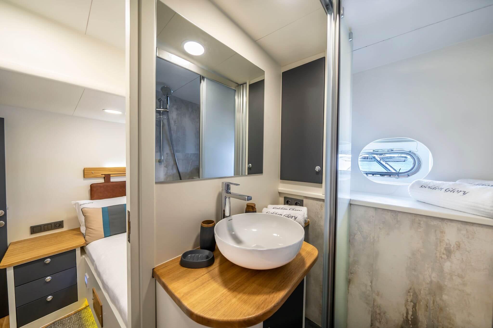 Golden Glory yacht en-suite bathroom with round vessel sink on teak vanity, overhead shower, and porthole window, adjacent to cabin bed