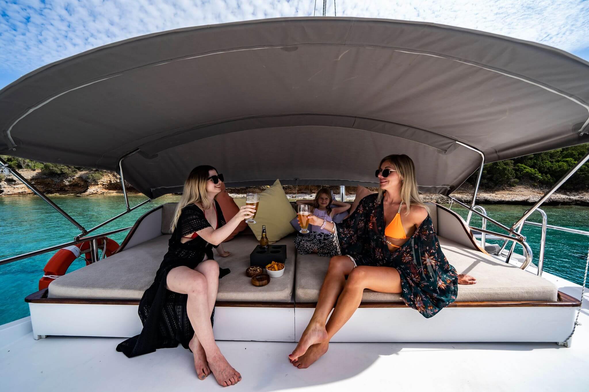 Two women clinking beer glasses on cushioned aft deck seating with snacks, child between them, turquoise cove backdrop