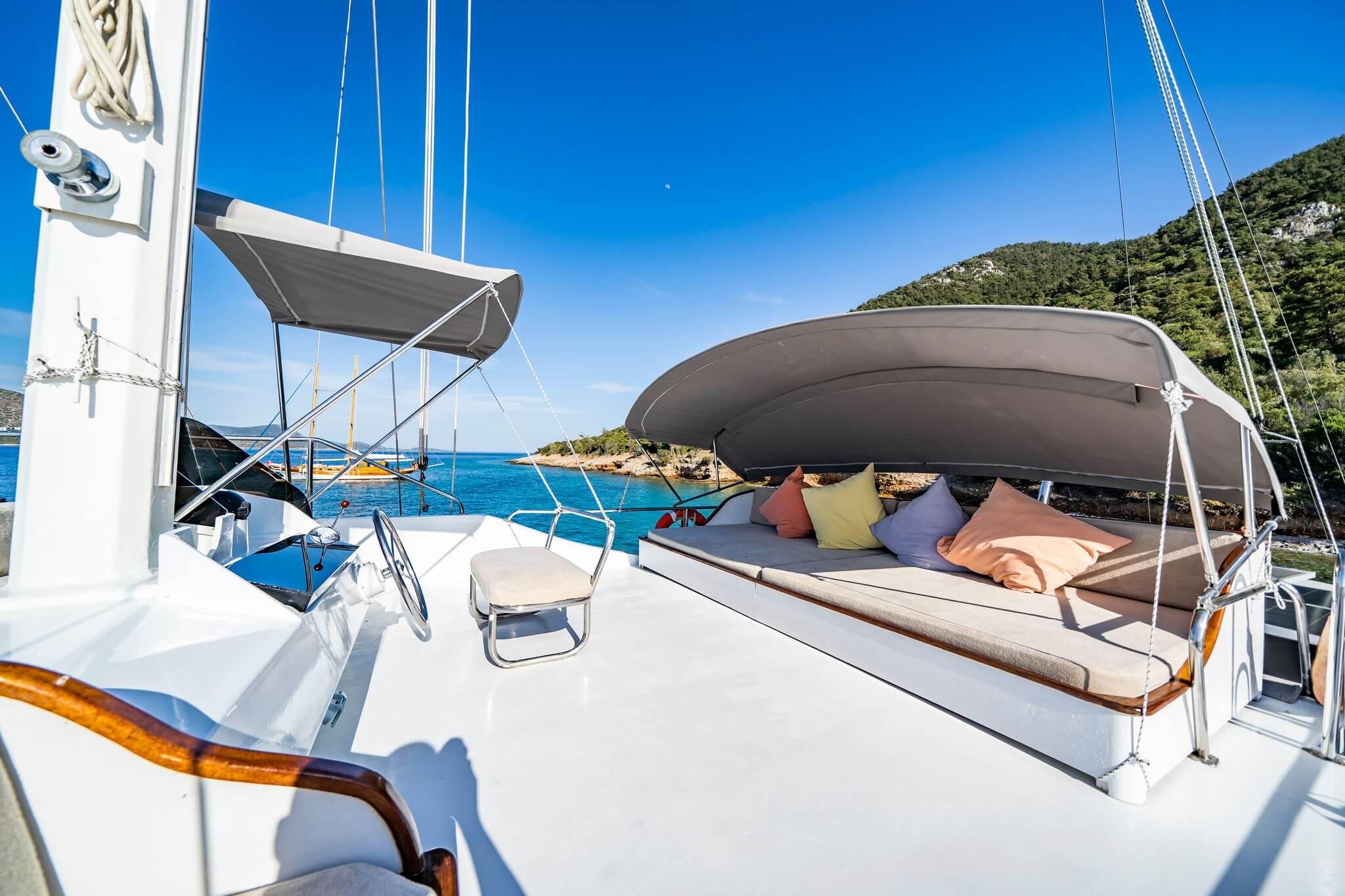 Gulet upper deck with stainless steel helm wheel, cushioned daybed with colourful scatter cushions under grey canopy, anchored in turquoise bay