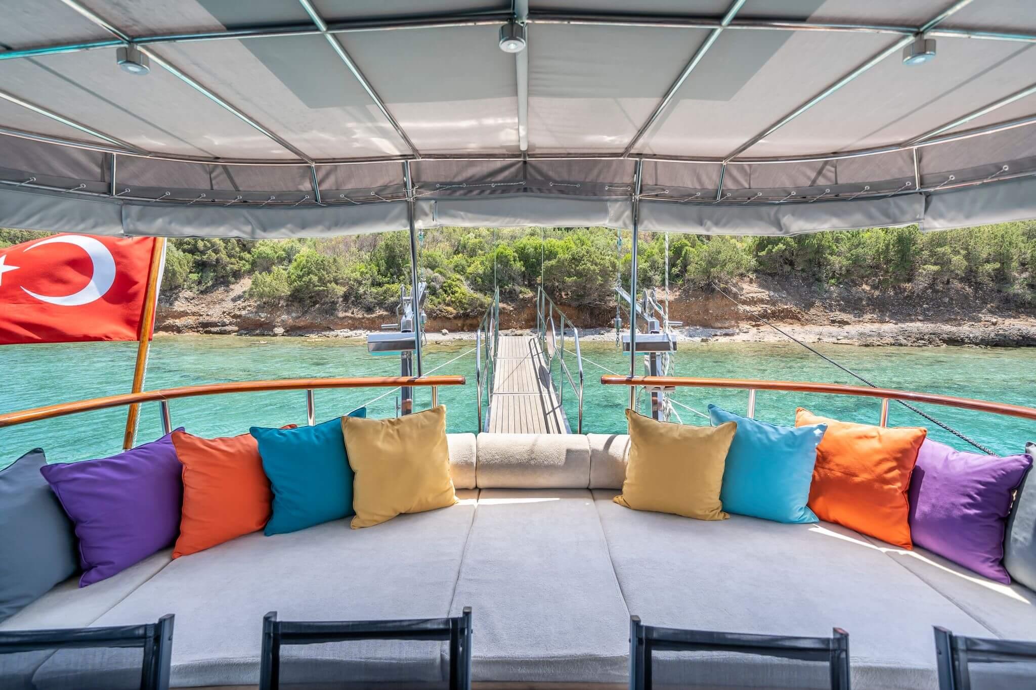 gulet aft deck curved sofa with colourful cushions, retractable canopy, and teak gangway leading to rocky shoreline