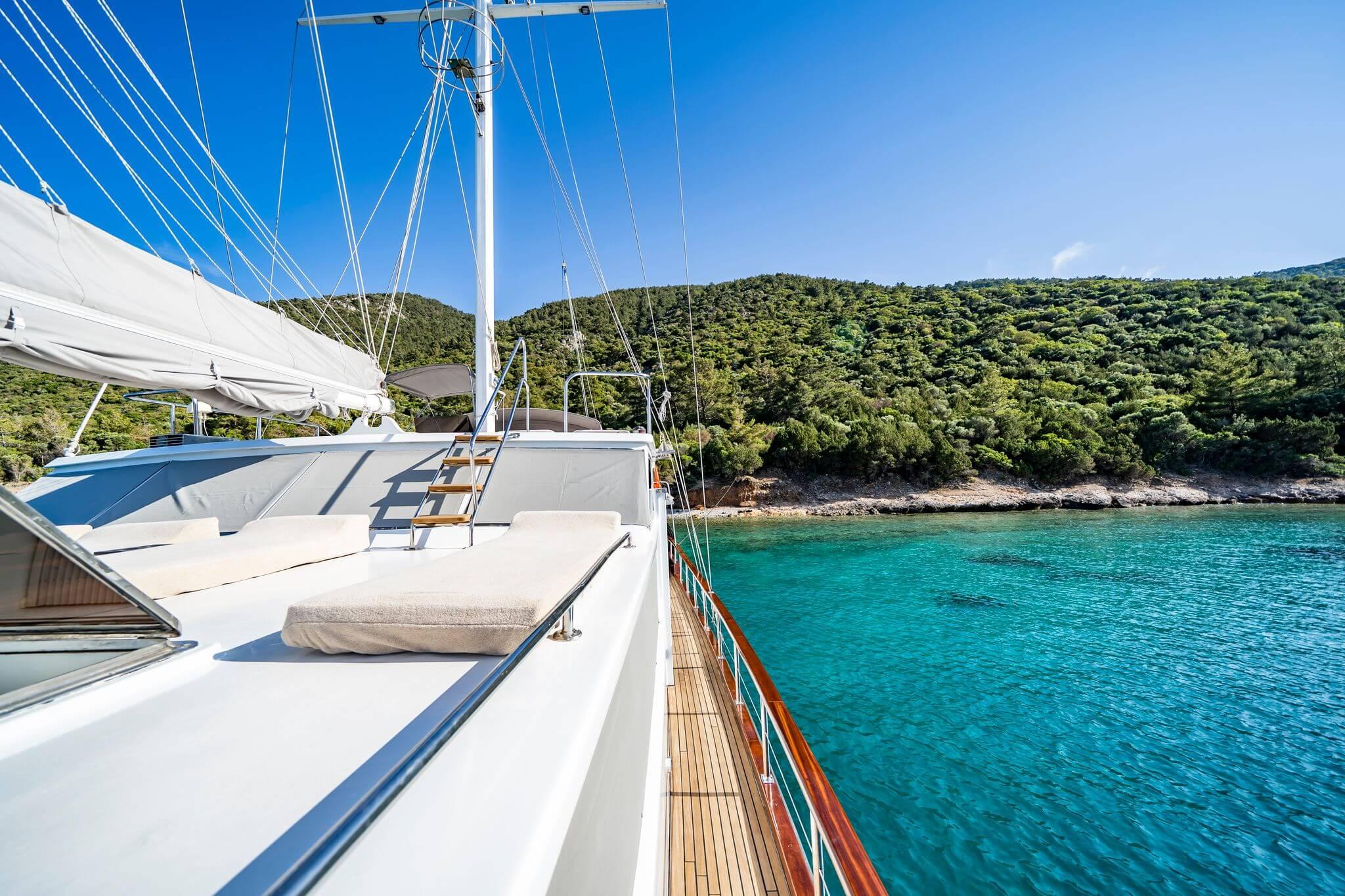 Luxury gulet side deck with white cushioned sun loungers, teak walkway, and turquoise bay anchorage