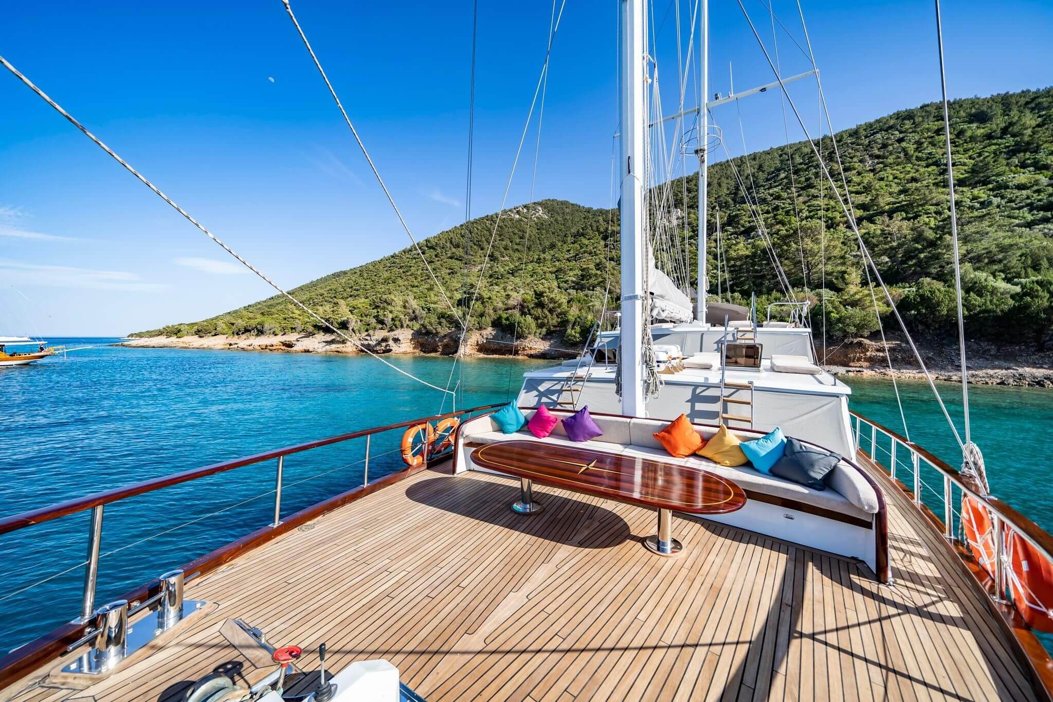 gulet bow deck with white upholstered sofa, colourful scatter cushions, oval teak coffee table, and turquoise bay anchorage
